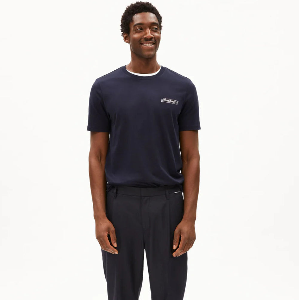 Ein Mann steht vor einem schlichten weißen Hintergrund und trägt ein marineblaues T-Shirt mit einem kleinen Logo auf der linken Brust, eine passende marineblaue Hose und weiße Turnschuhe. Er lächelt und blickt leicht zur Seite.