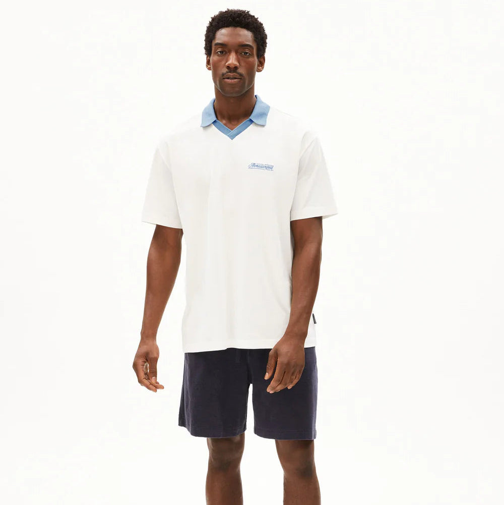 Ein Mann steht in einem Studio und trägt ein weißes kurzärmeliges Polohemd mit hellblauem Kragen, marineblaue Shorts und hellblaue Turnschuhe vor einem schlichten weißen Hintergrund.