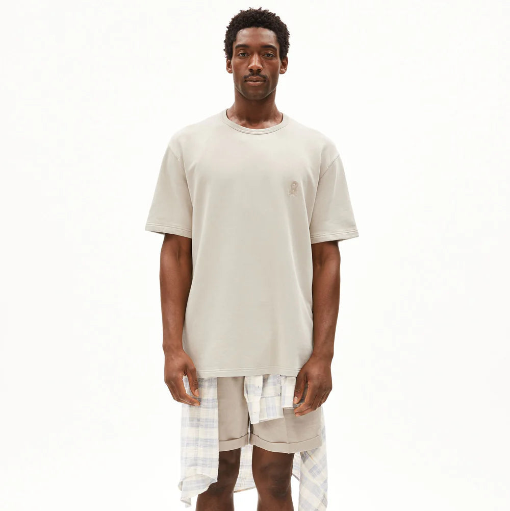 Ein Mann steht mit dem Gesicht nach vorne und trägt ein beigefarbenes T-Shirt, beigefarbene Shorts mit einem um die Taille gebundenen karierten Hemd und schwarze Sandalen vor einem schlichten weißen Hintergrund.