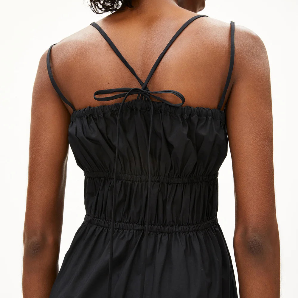 Eine Person mit dunklem, gelocktem Haar wird von hinten gezeigt. Sie trägt ein schwarzes Spaghettiträger-Kleid mit elastischen Raffungen und dünnen Trägern, die im oberen Rückenbereich zu einer Schleife gebunden sind.