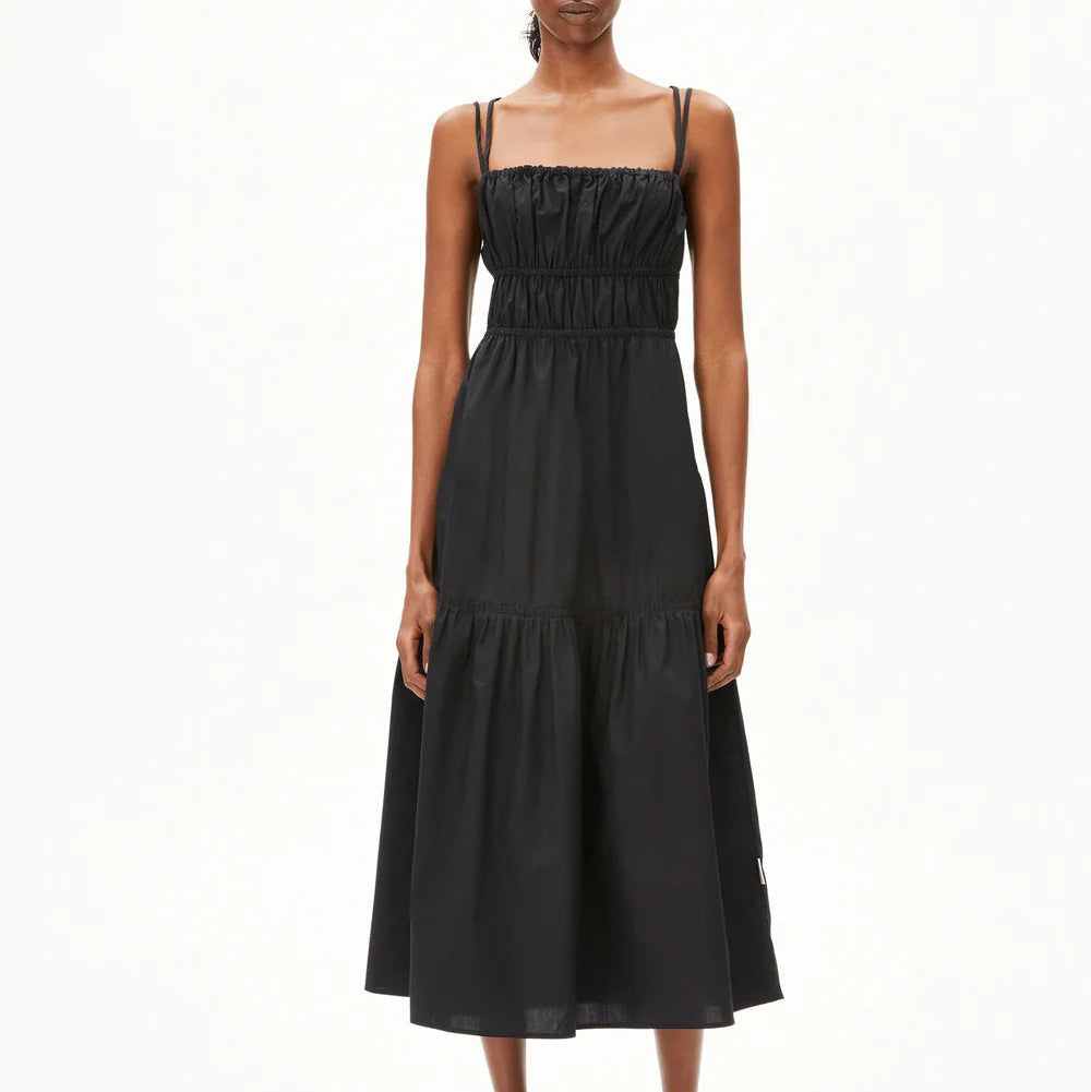 Eine Frau steht mit dem Gesicht nach vorne und trägt ein ärmelloses, schwarzes, knöchellanges Kleid mit dünnen Trägern und Raffungen an Mieder und Taille. Sie trägt außerdem schwarze flache Sandalen. Der Hintergrund ist einfarbig weiß.