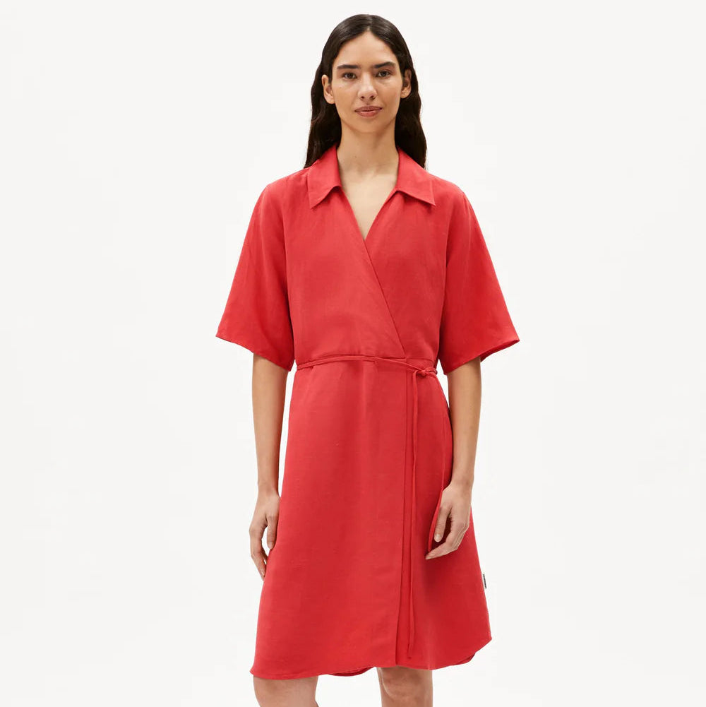 Eine Frau steht vor einem weißen Hintergrund und trägt ein knielanges, kurzärmeliges rotes Wickelkleid mit Kragen, dazu schwarze, zehenoffene Slide-Sandalen. Sie hat langes braunes Haar und einen neutralen Gesichtsausdruck.