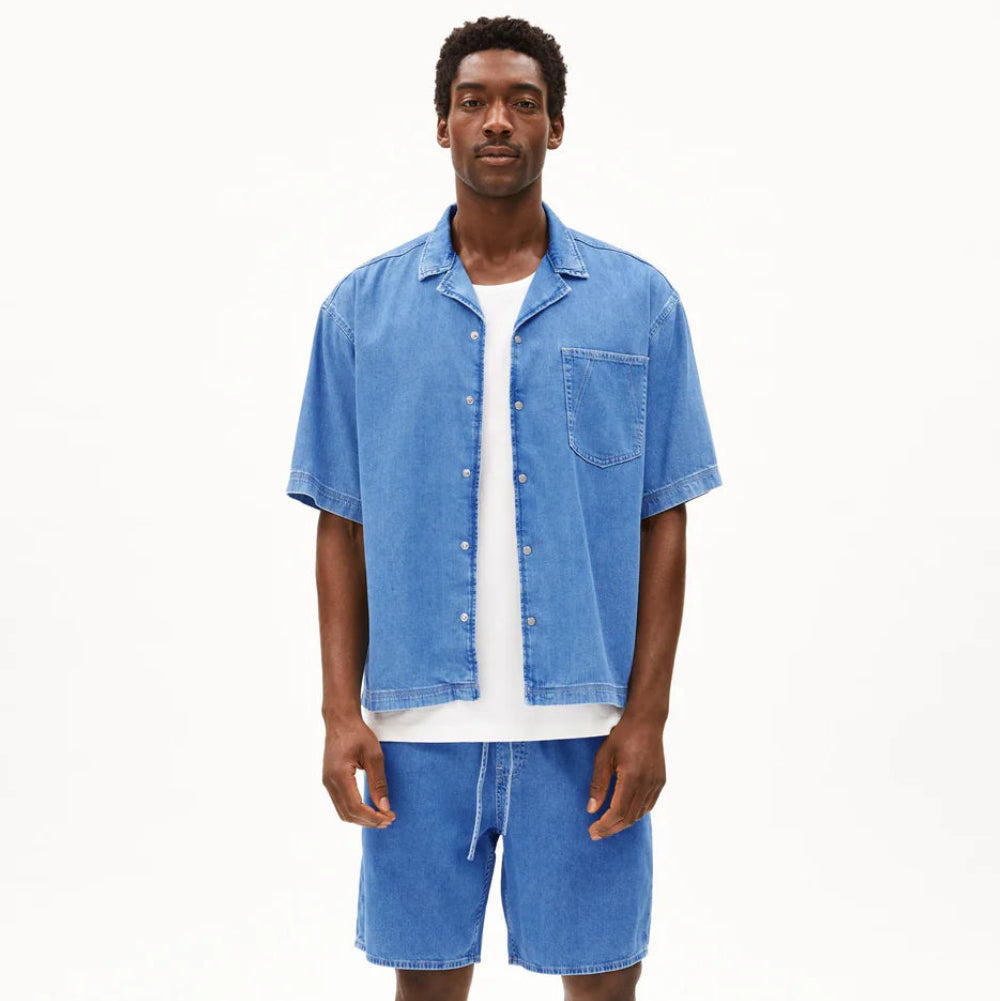 Ein Mann steht mit dem Gesicht nach vorne und trägt ein kurzärmeliges Jeanshemd mit Knopfleiste über einem weißen T-Shirt, blaue Shorts mit Kordelzug und schwarze Sandalen mit Korksohlen vor einem schlichten weißen Hintergrund.