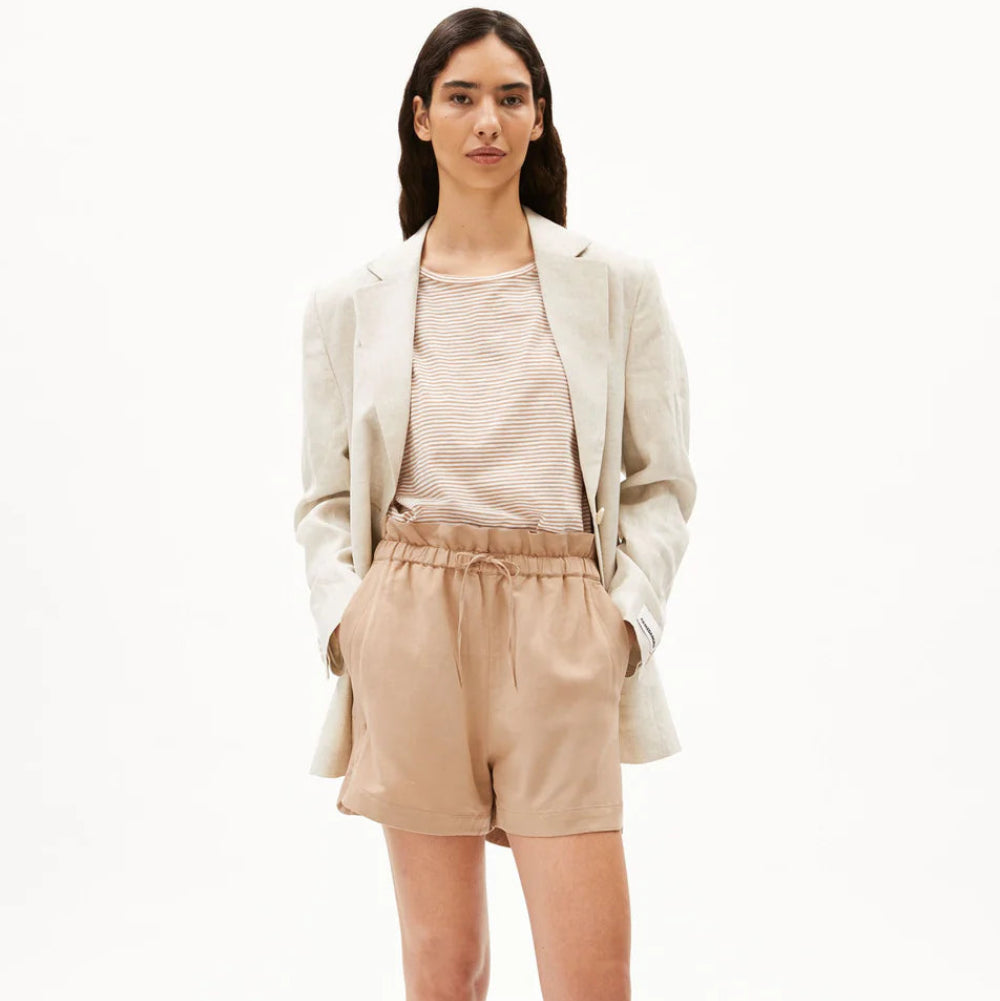 Eine Person steht vor einem weißen Hintergrund und trägt einen hellbeigen Blazer, ein beiges T-Shirt, hellbraune Shorts mit Kordelzug und schwarze Sandalen mit grünen Details. Ihre Hände stecken in den Taschen und sie hat langes dunkles Haar.