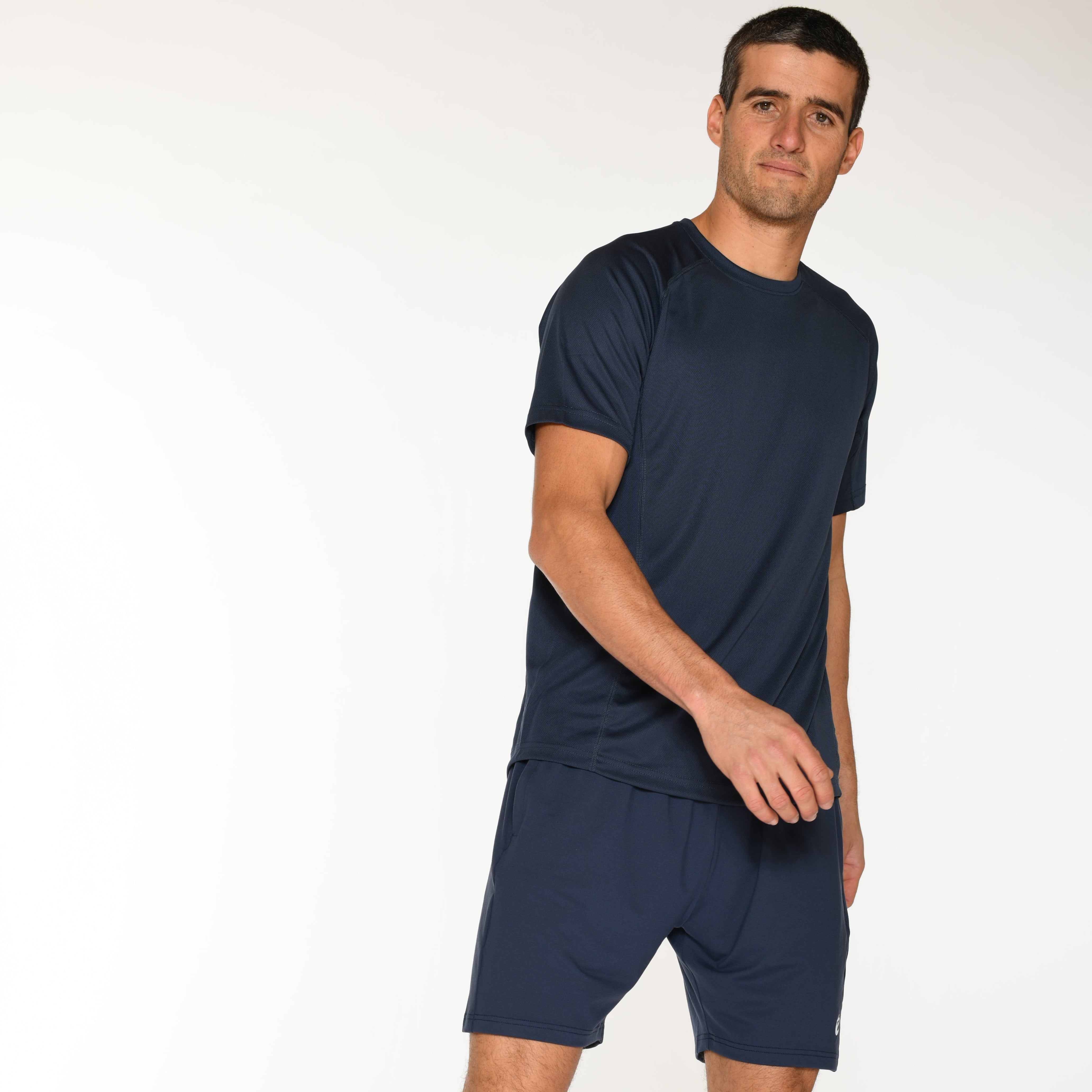 Ein Mann in marineblauem Sportdress und blauen Turnschuhen geht vor einem schlichten weißen Hintergrund nach vorne und schaut leicht nach rechts.