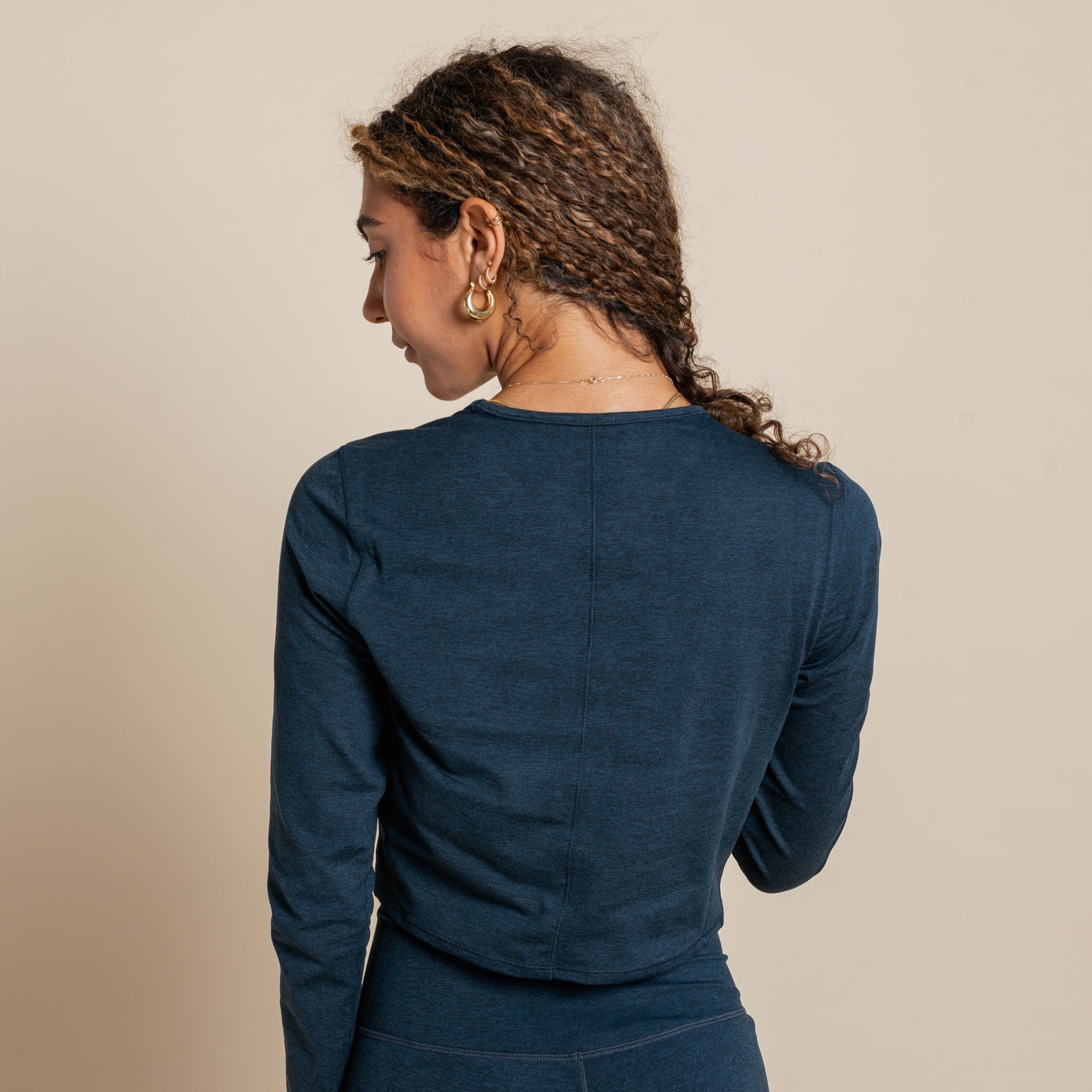 Frau mit lockigem Haar und blauen Oberteil steht mit dem Rücken zur Kamera vor einem beigen Hintergrund.