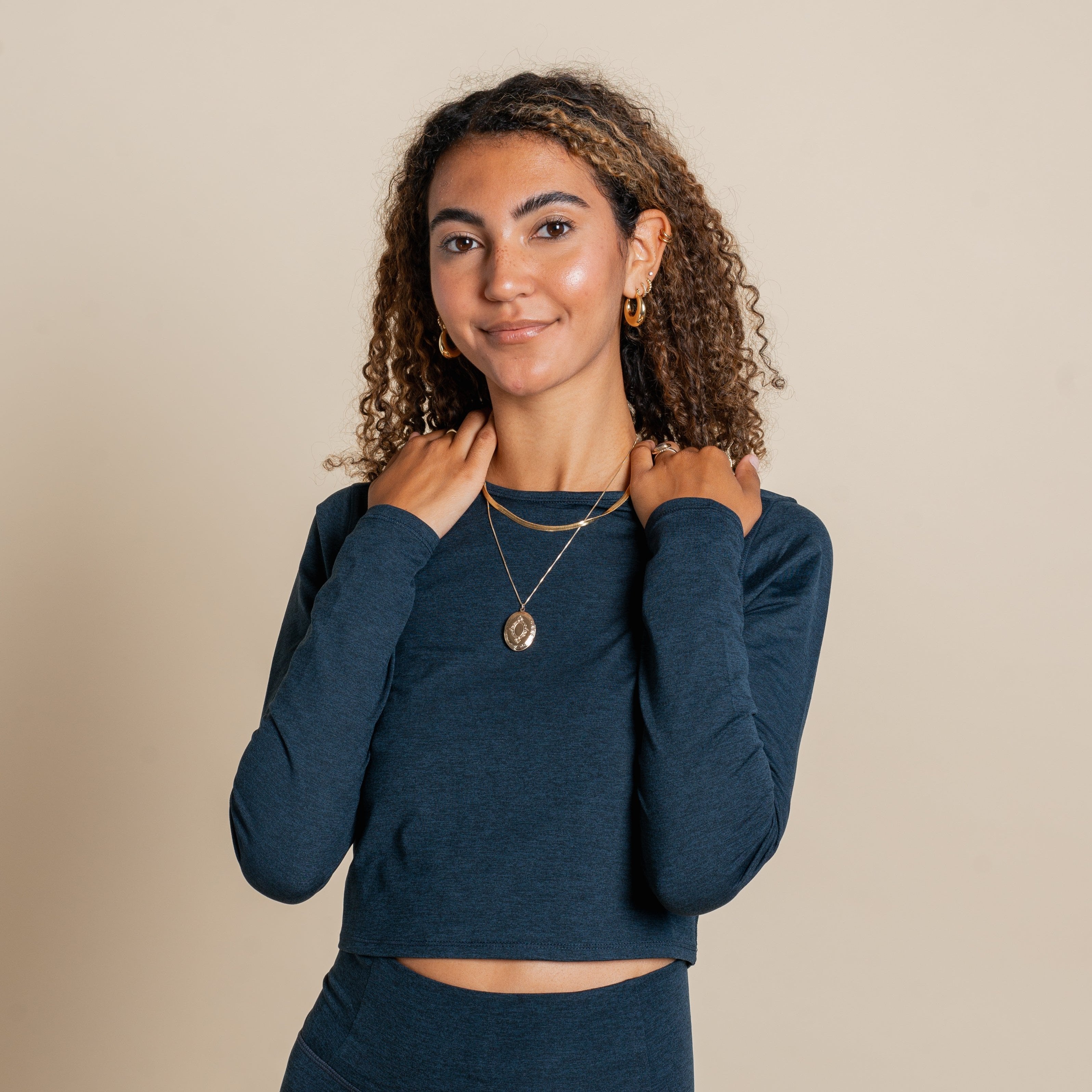 Frau in dunkelblauem Shirt mit goldenen Ohrringen und Ketten vor beigem Hintergrund lächelt in die Kamera.