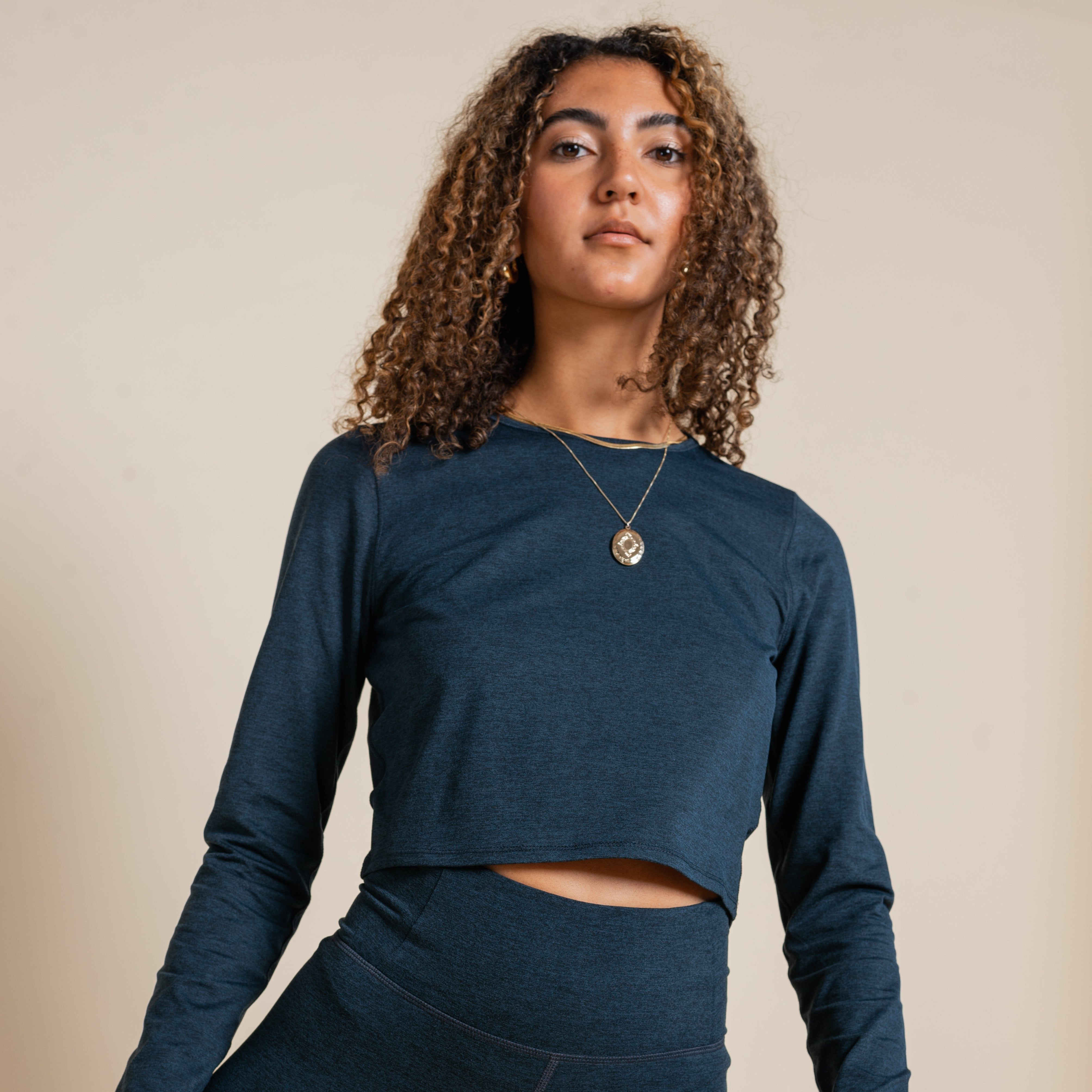 Eine Frau mit lockigem Haar trägt ein dunkelblaues, langärmliges Oberteil und eine Halskette vor neutralem Hintergrund.