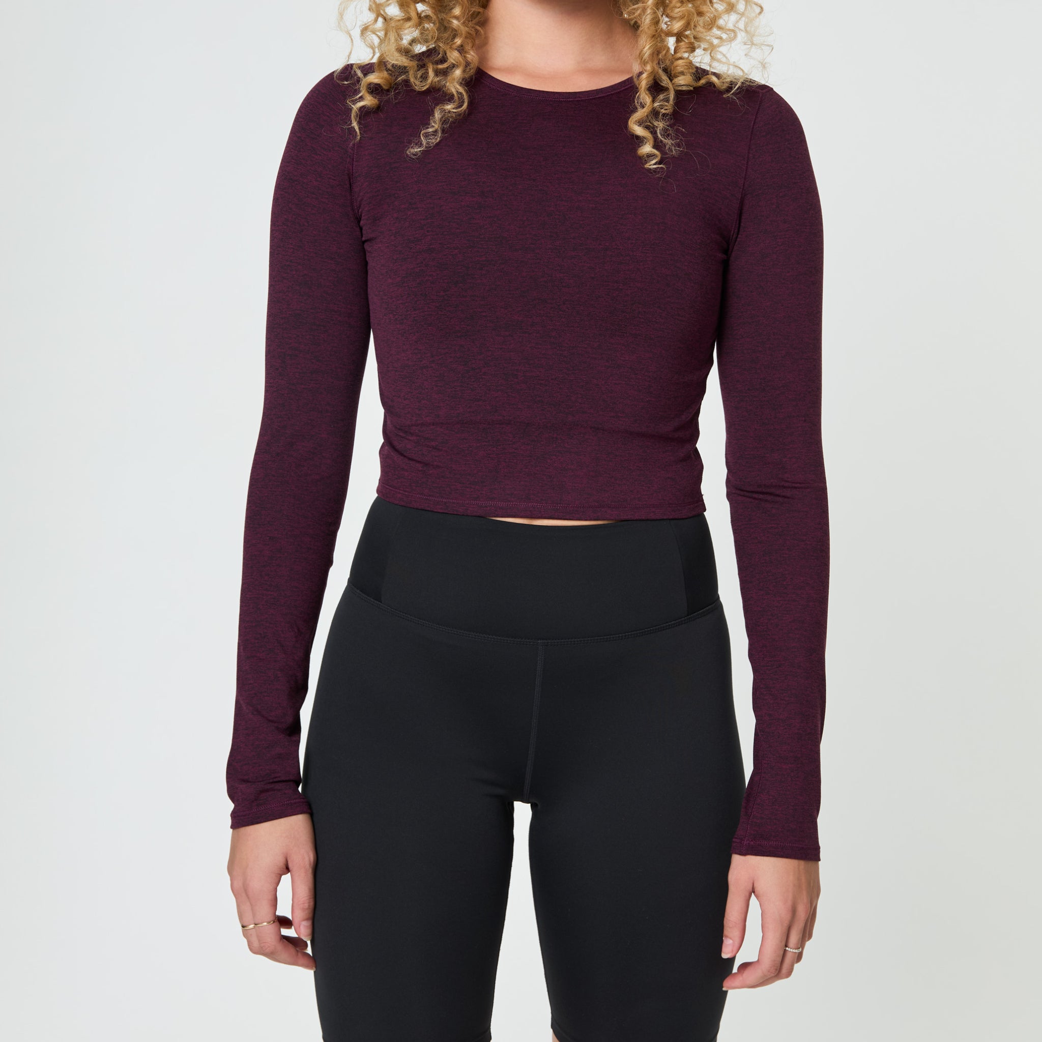 Person in burgundy long-sleeve top and black high-waisted leggings, with curly hair, standing against a plain background.
