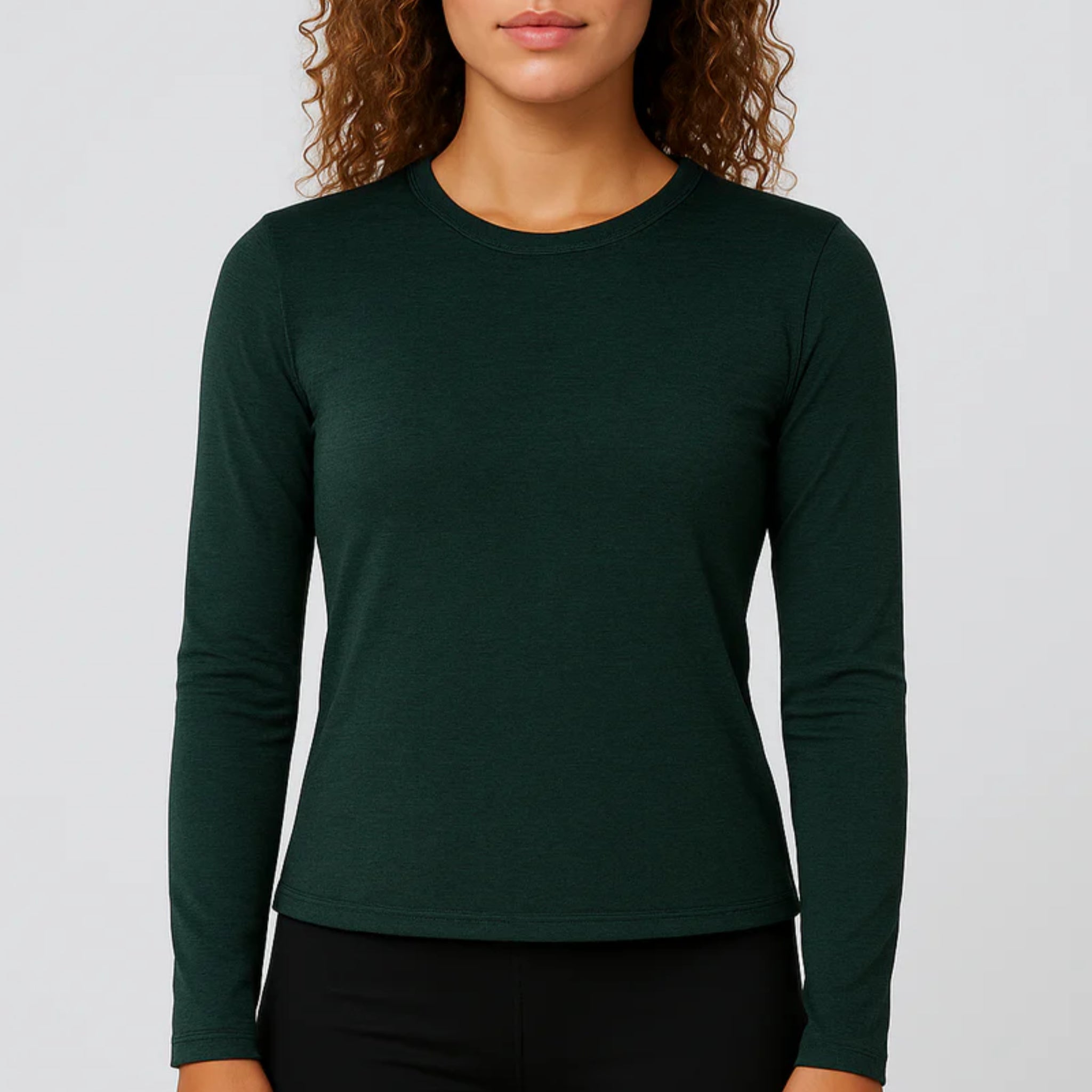 Eine Frau mit lockigem Haar trägt ein dunkelgrünes, langärmeliges T-Shirt mit Rundhalsausschnitt und eine schwarze Hose und steht vor einem schlichten hellgrauen Hintergrund.