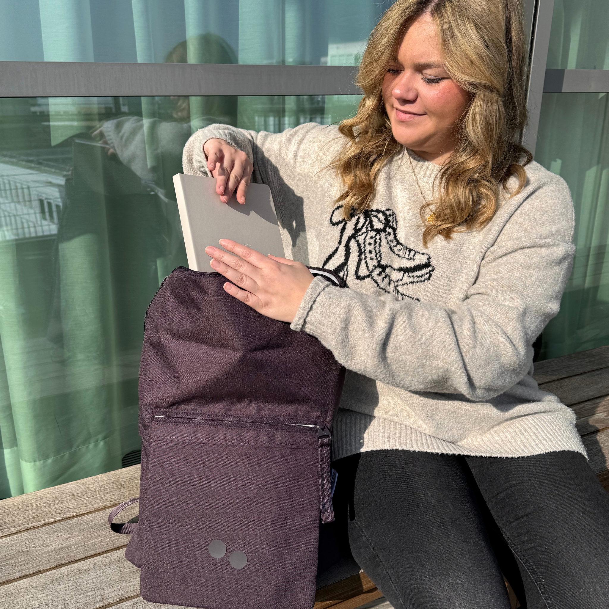 Frau mit langen Haaren steckt ein Buch in einen lila Rucksack, während sie auf einer Bank in der Sonne sitzt.