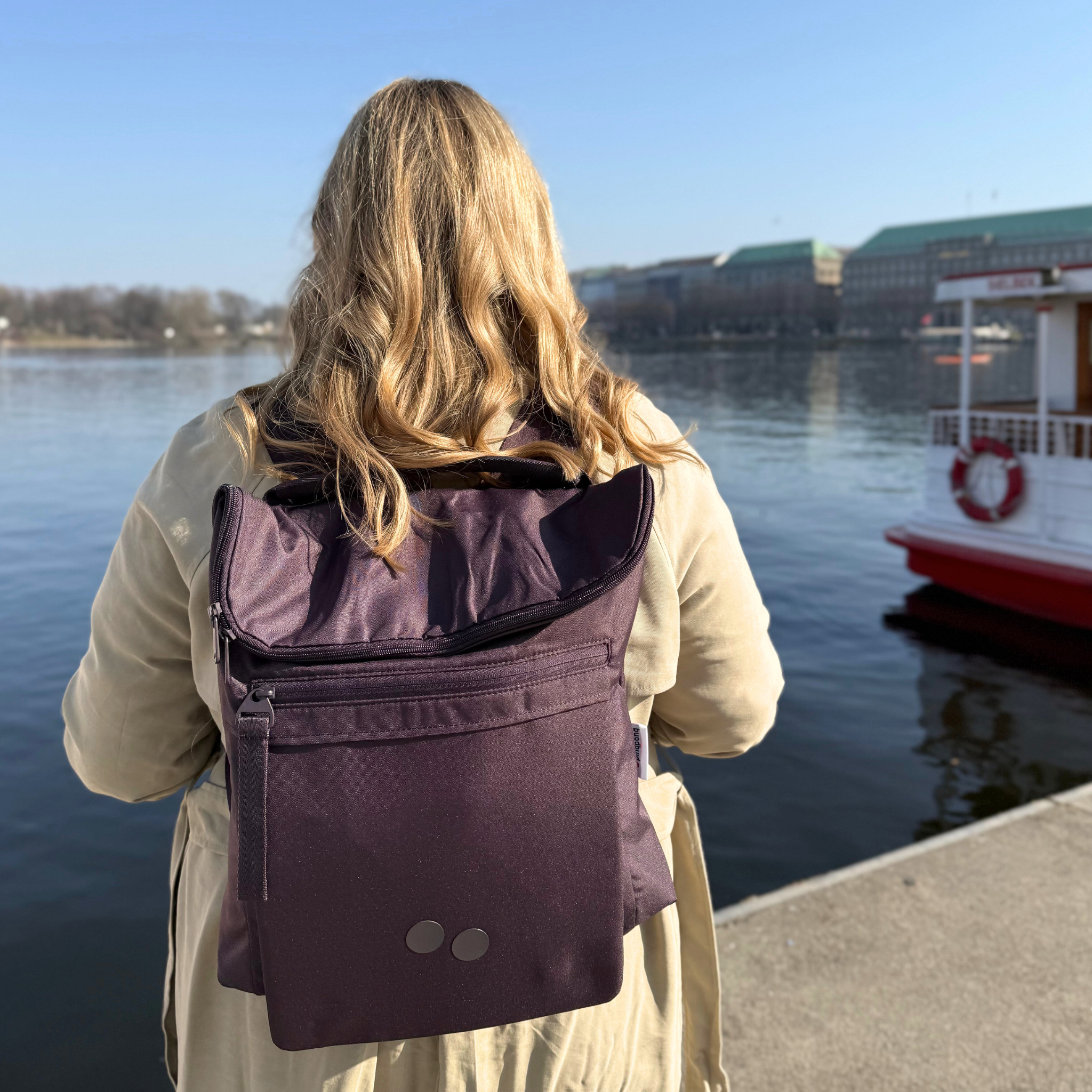 Person mit lilafarbenem Rucksack blickt auf einen Fluss, im Hintergrund ist ein weißes Boot zu sehen.
