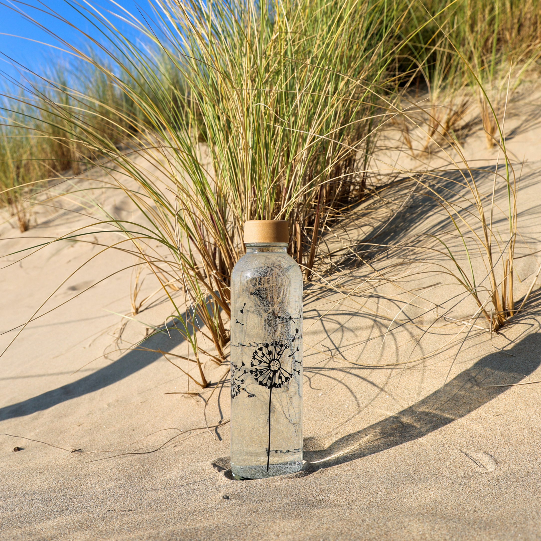Eine Glasflasche mit Wasser und Löwenzahnmotiv steht auf einem Sandstrand neben Grashalmen.