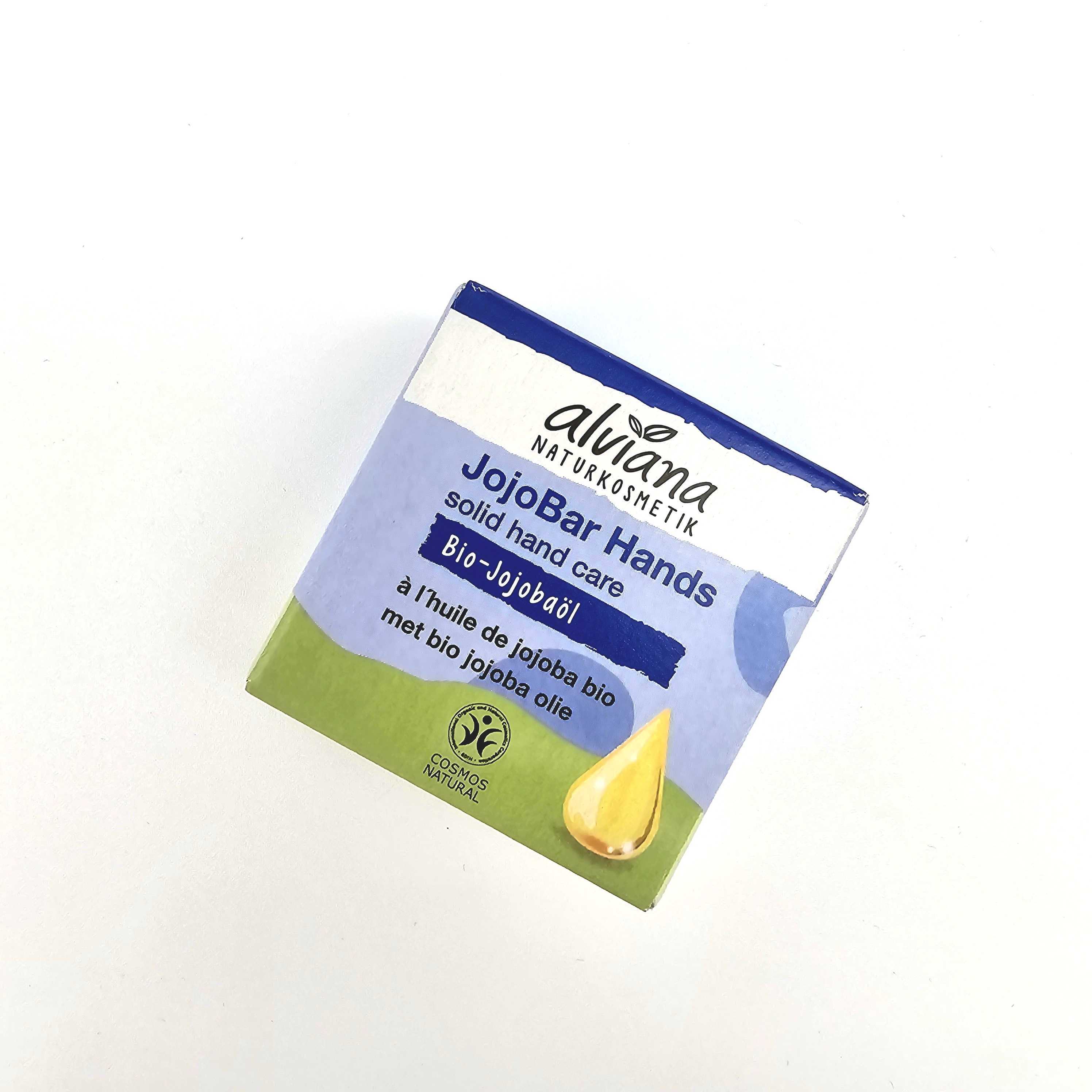 Eine quadratische Dose alviana NATURKOSMETIK JojobaBar Hands solide Handpflege, mit blauen, weißen und grünen Farben, einem gelben Jojobaöltropfen und Text in Englisch, Deutsch und Französisch.