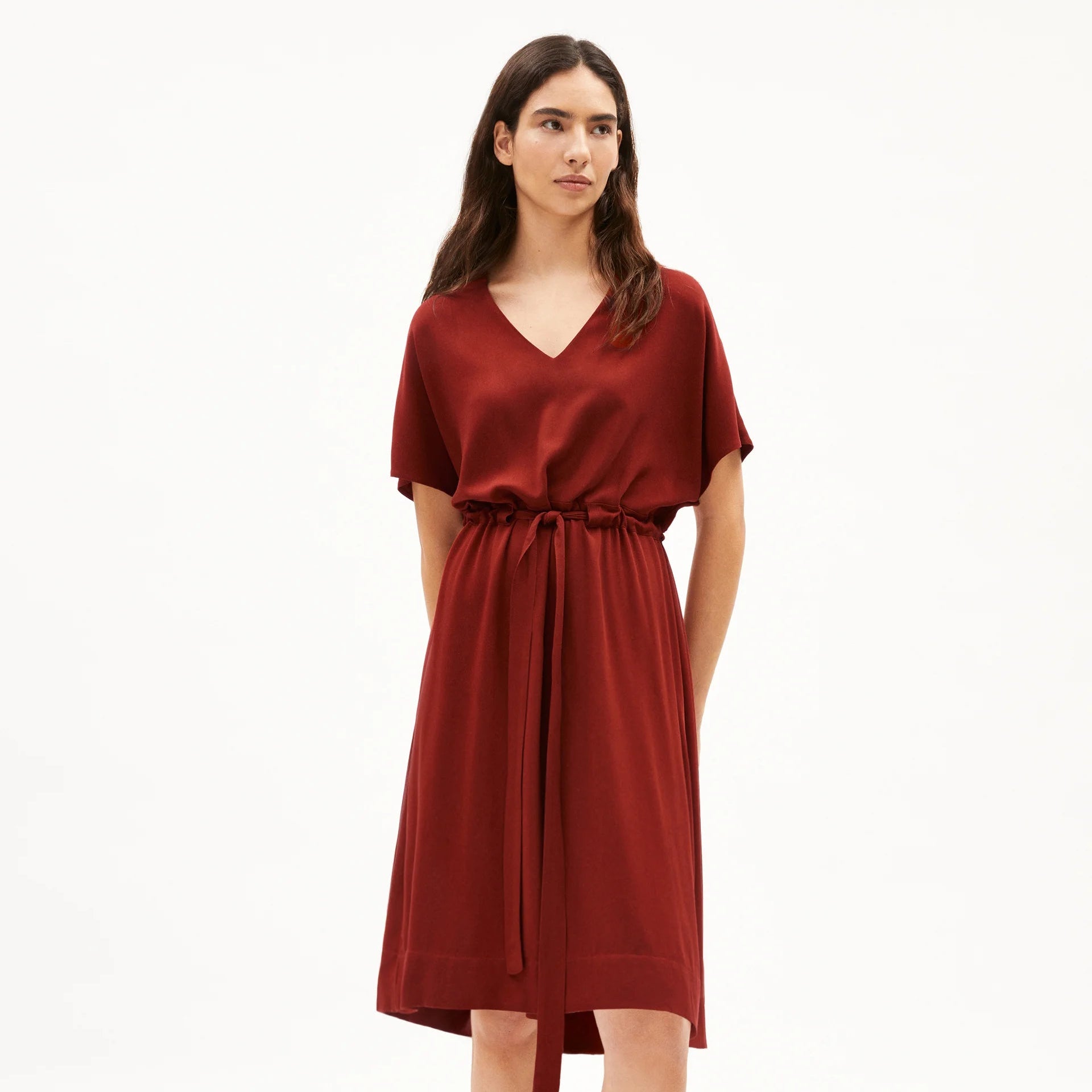 Eine Frau trägt ein kurzärmeliges, knielanges, rotes Kleid mit V-Ausschnitt und Bindegürtel vor einem schlichten weißen Hintergrund.