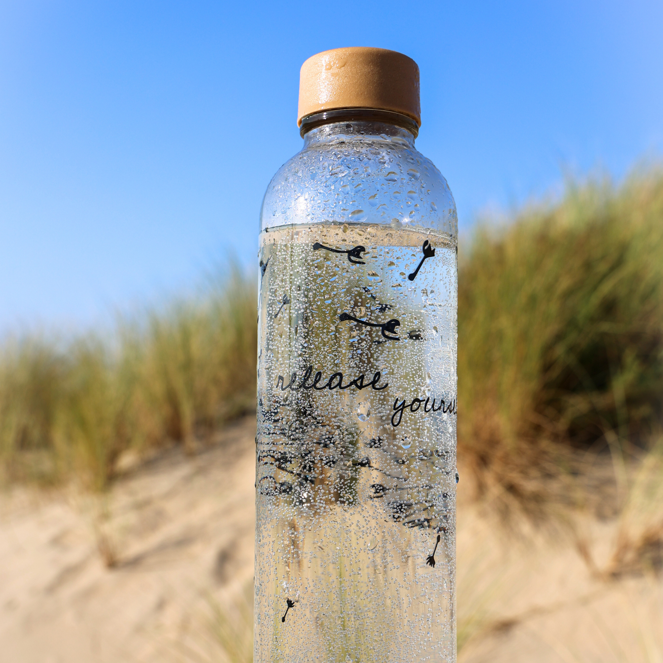 Glasflasche mit Wasser und Korkverschluss vor unscharfem Dünenhintergrund unter klarem blauem Himmel.