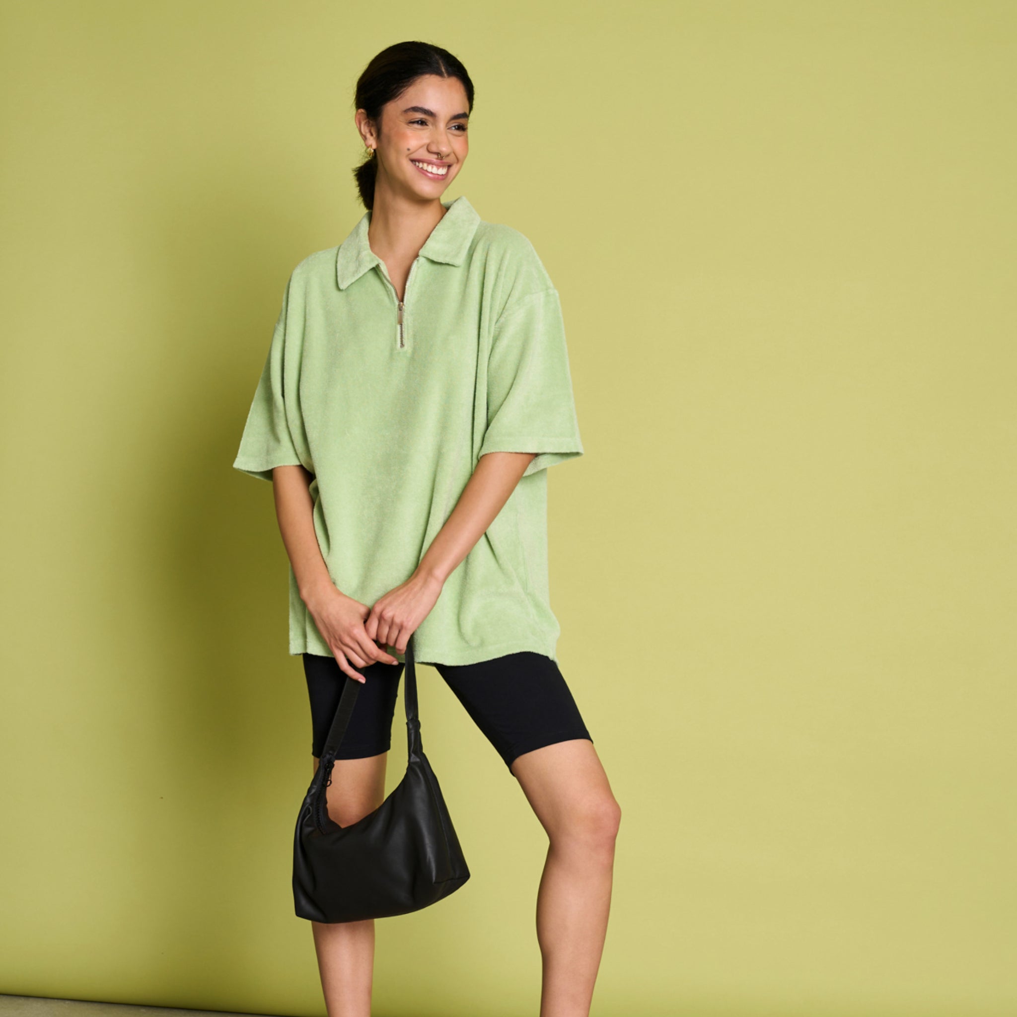 Frau in mintgrünem Shirt und schwarzer Shorts hält eine schwarze Tasche vor einem hellgrünen Hintergrund.