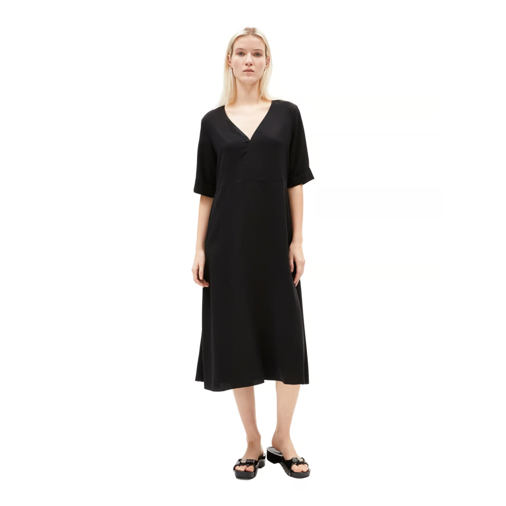 Eine Frau steht vor einem weißen Hintergrund und trägt ein locker sitzendes, knielanges schwarzes Kleid mit kurzen Ärmeln und einem V-Ausschnitt, dazu schwarze Sandalen.