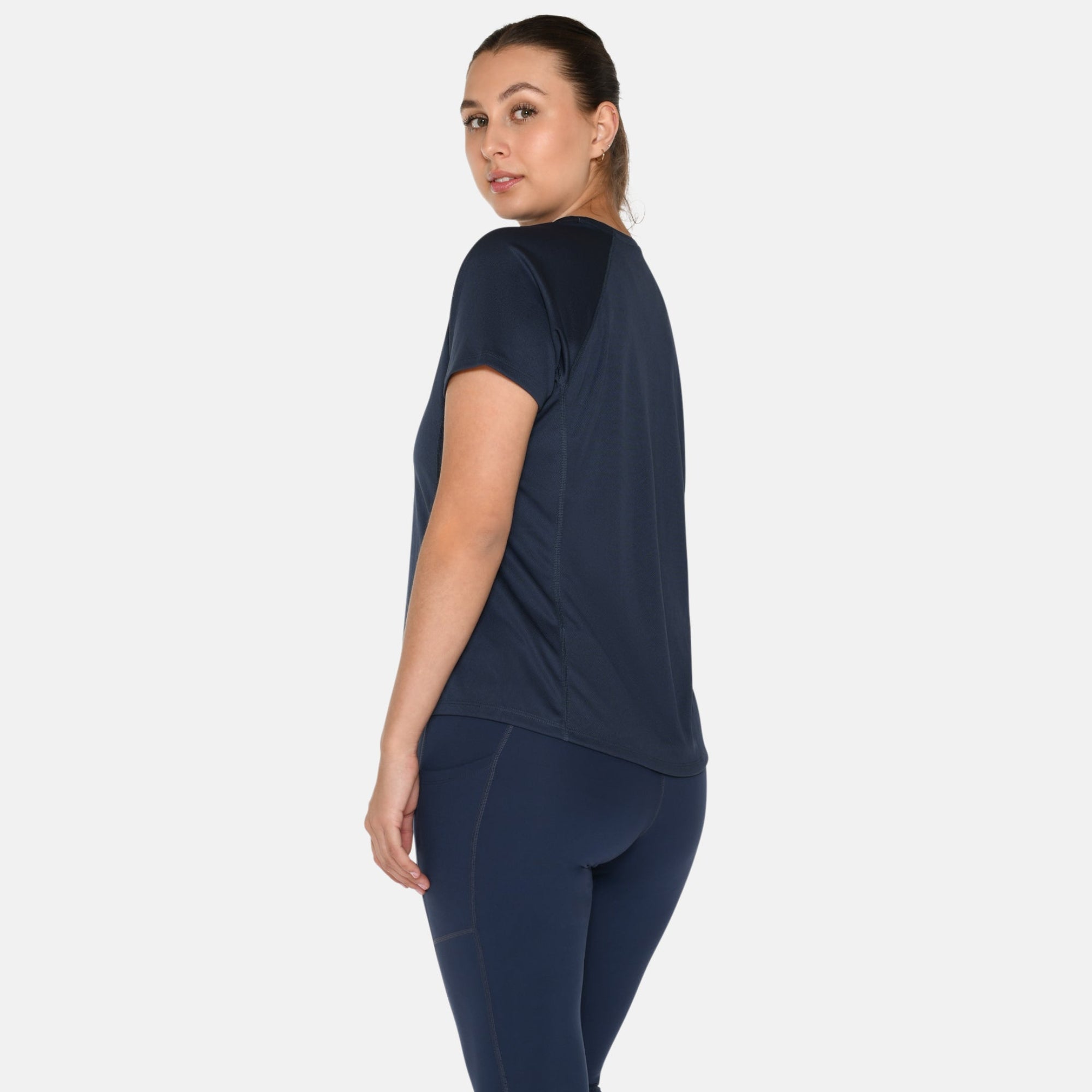 Eine Frau, die ein marineblaues Sport-T-Shirt und passende Leggings trägt, steht leicht rückwärtsgewandt und schaut über ihre Schulter. Sie trägt weiße Turnschuhe und steht vor einem schlichten weißen Hintergrund.