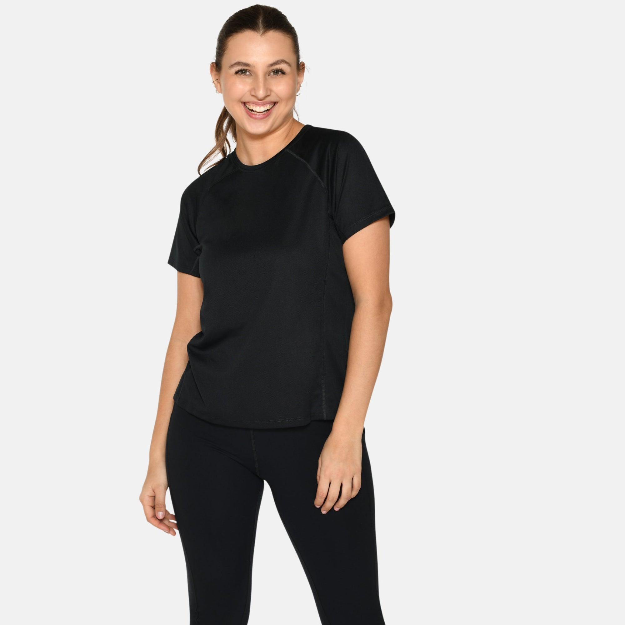 Eine Frau steht lächelnd vor einem schlichten Hintergrund und trägt ein schwarzes kurzärmeliges Oberteil, schwarze Leggings und weiße Turnschuhe.