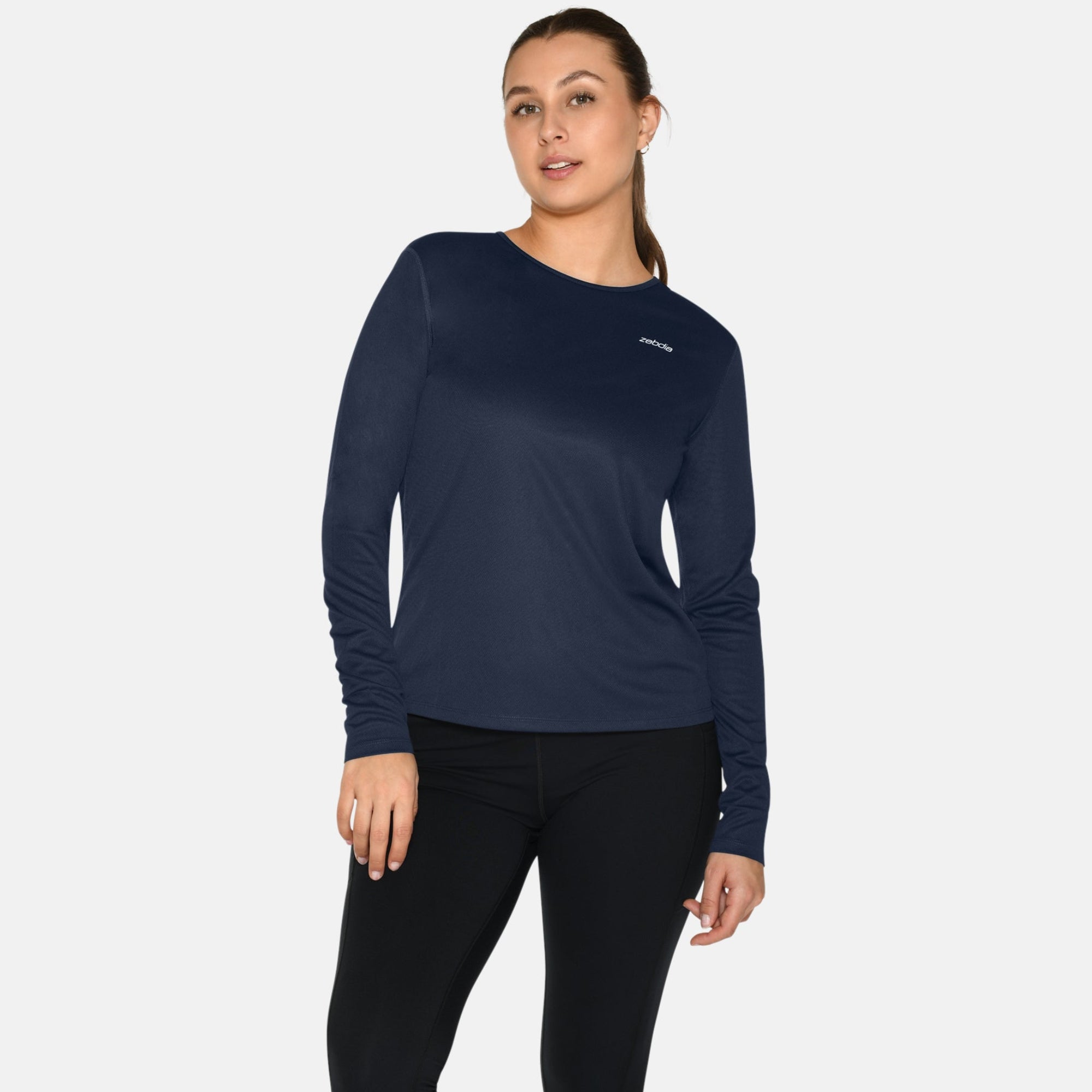 Eine Frau steht vor einem weißen Hintergrund und trägt ein marineblaues langärmeliges Hemd, schwarze Leggings und weiße Turnschuhe. Sie hat ihr Haar zurückgebunden und blickt mit entspanntem Gesichtsausdruck und lässiger Haltung nach vorne.