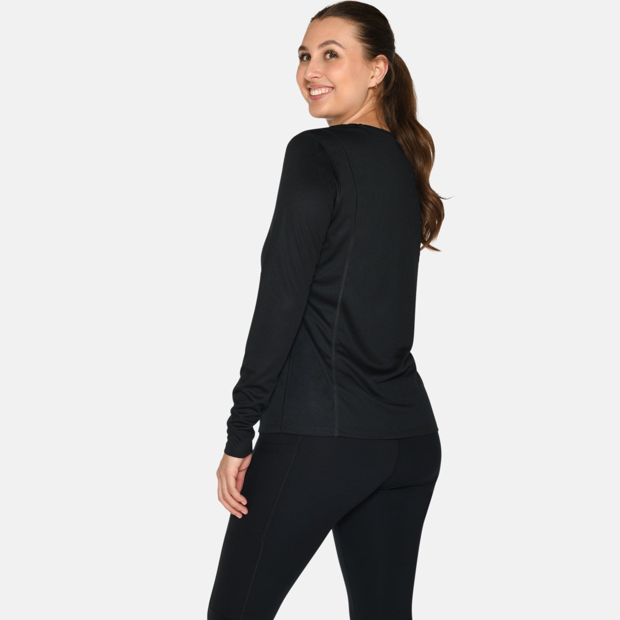 Eine Frau mit langen braunen Haaren in einem Pferdeschwanz trägt ein schwarzes langärmeliges Oberteil, schwarze Leggings und weiße Turnschuhe. Sie steht und lächelt, leicht von der Kamera abgewandt, vor einem weißen Hintergrund.