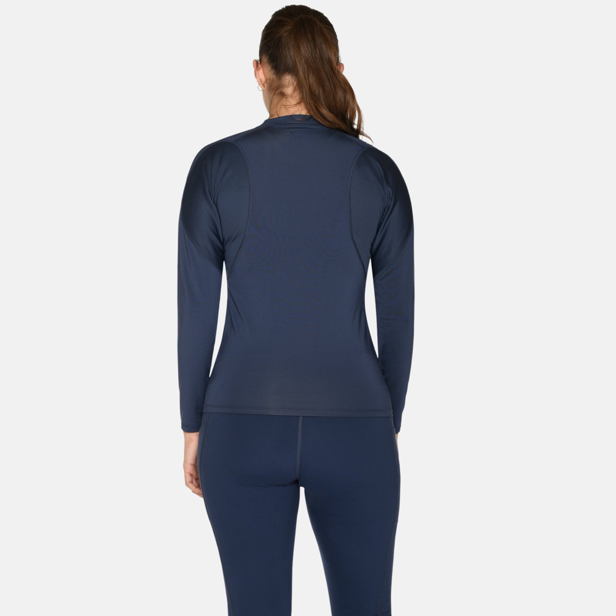 Eine Frau steht mit dem Gesicht zur Kamera, trägt ein marineblaues langärmeliges Sportoberteil, passende Leggings und weiße Turnschuhe vor einem schlichten weißen Hintergrund.