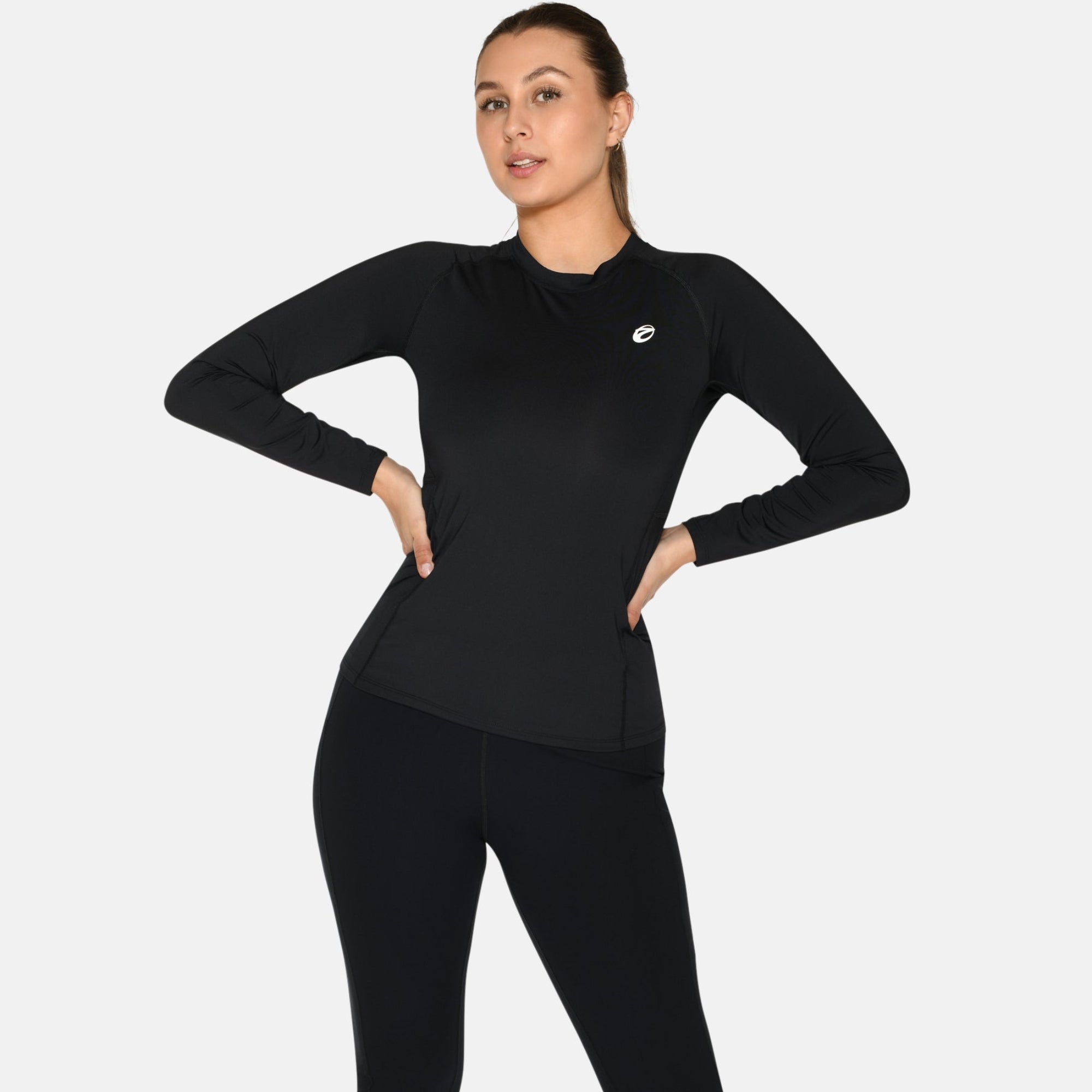 Eine Person steht selbstbewusst mit den Händen in den Hüften, trägt ein schwarzes langärmeliges Sportoberteil, schwarze Leggings und weiße Turnschuhe vor einem schlichten weißen Hintergrund.