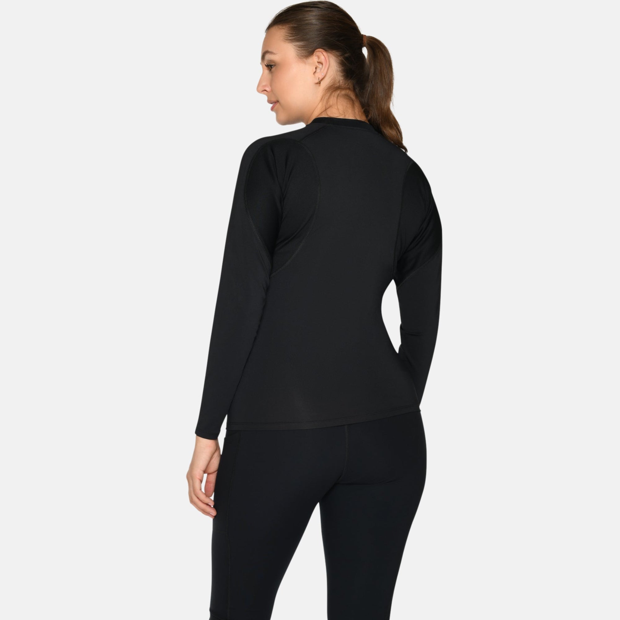 Eine Frau mit braunen Haaren in einem Pferdeschwanz steht und schaut weg. Sie trägt ein schwarzes langärmeliges Sportoberteil, schwarze Leggings und weiße Turnschuhe vor einem schlichten hellen Hintergrund.