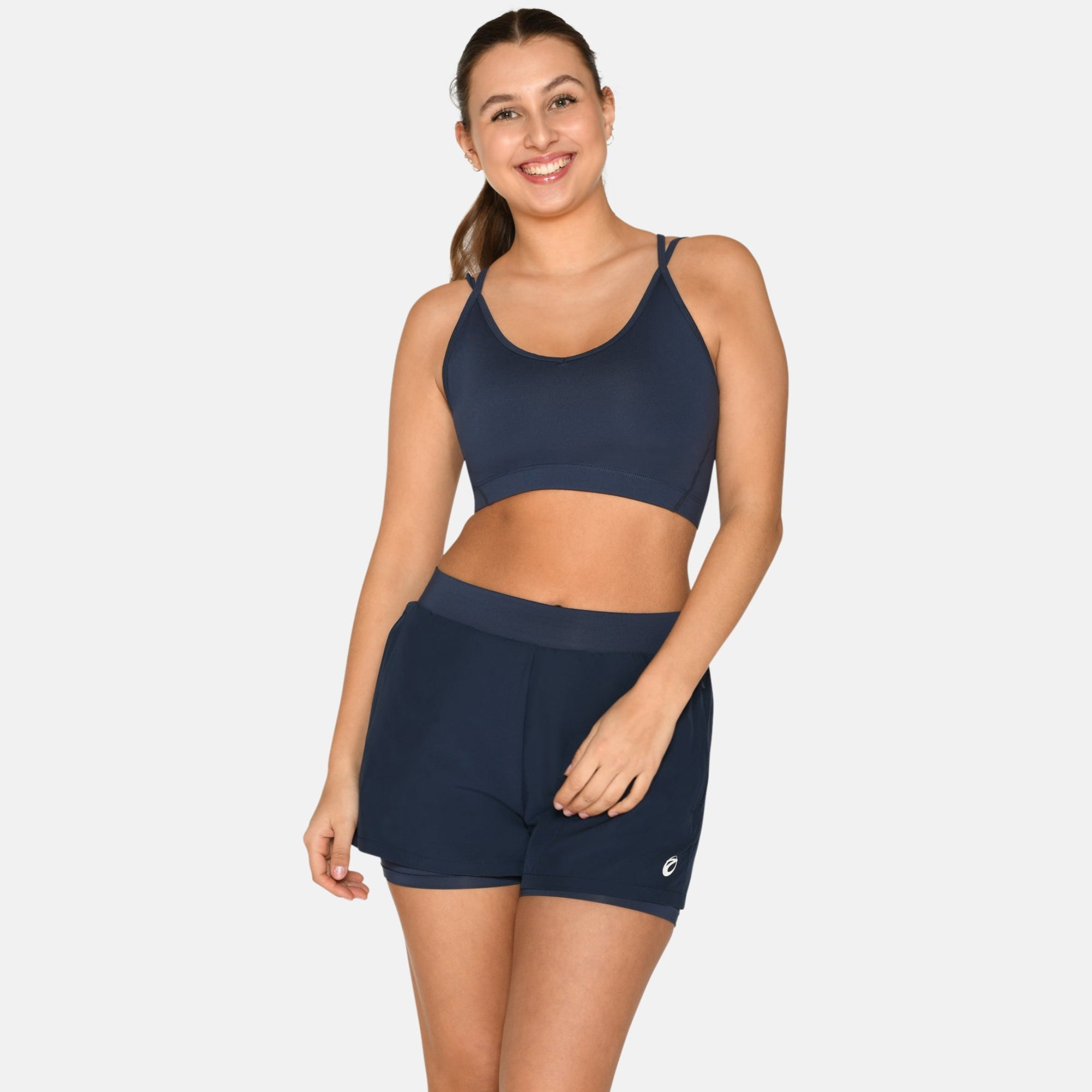 Eine Frau lächelt, während sie einen marineblauen Sport-BH, passende Shorts und weiße Turnschuhe trägt. Sie steht vor einem schlichten weißen Hintergrund, der ihr ein lässiges, sportliches Aussehen verleiht.