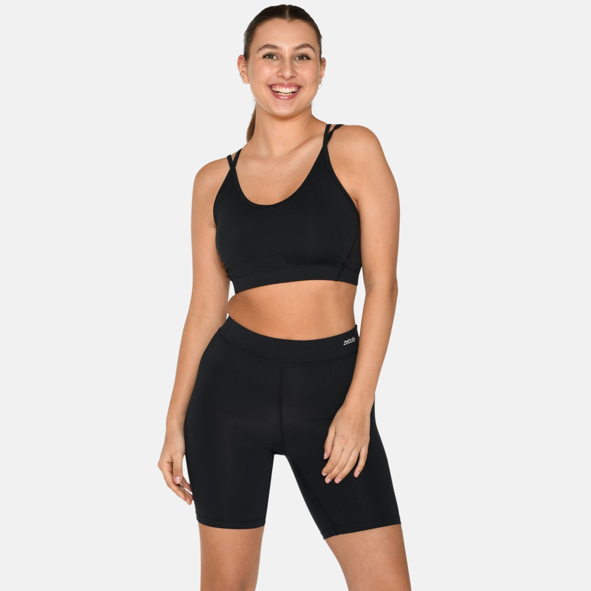 Eine lächelnde Frau steht vor einem schlichten Hintergrund und trägt einen schwarzen Sport-BH, schwarze, hoch taillierte Radlerhosen und weiße Turnschuhe. Sie hat ihr Haar zurückgebunden und die Hände an den Seiten.