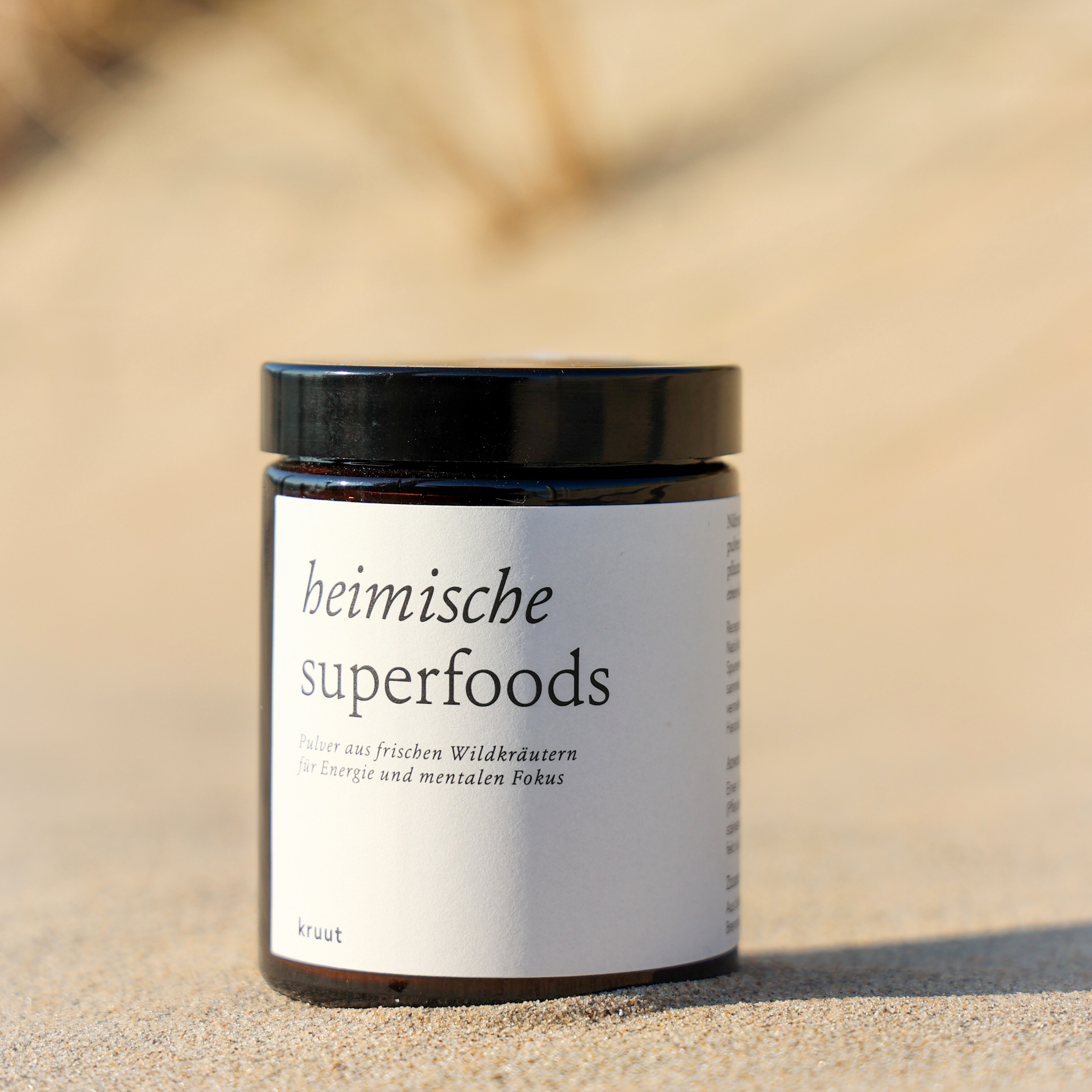 Braunes Glas mit schwarzem Deckel, auf dem Etikett steht "heimische superfoods" in einer sandigen Umgebung.