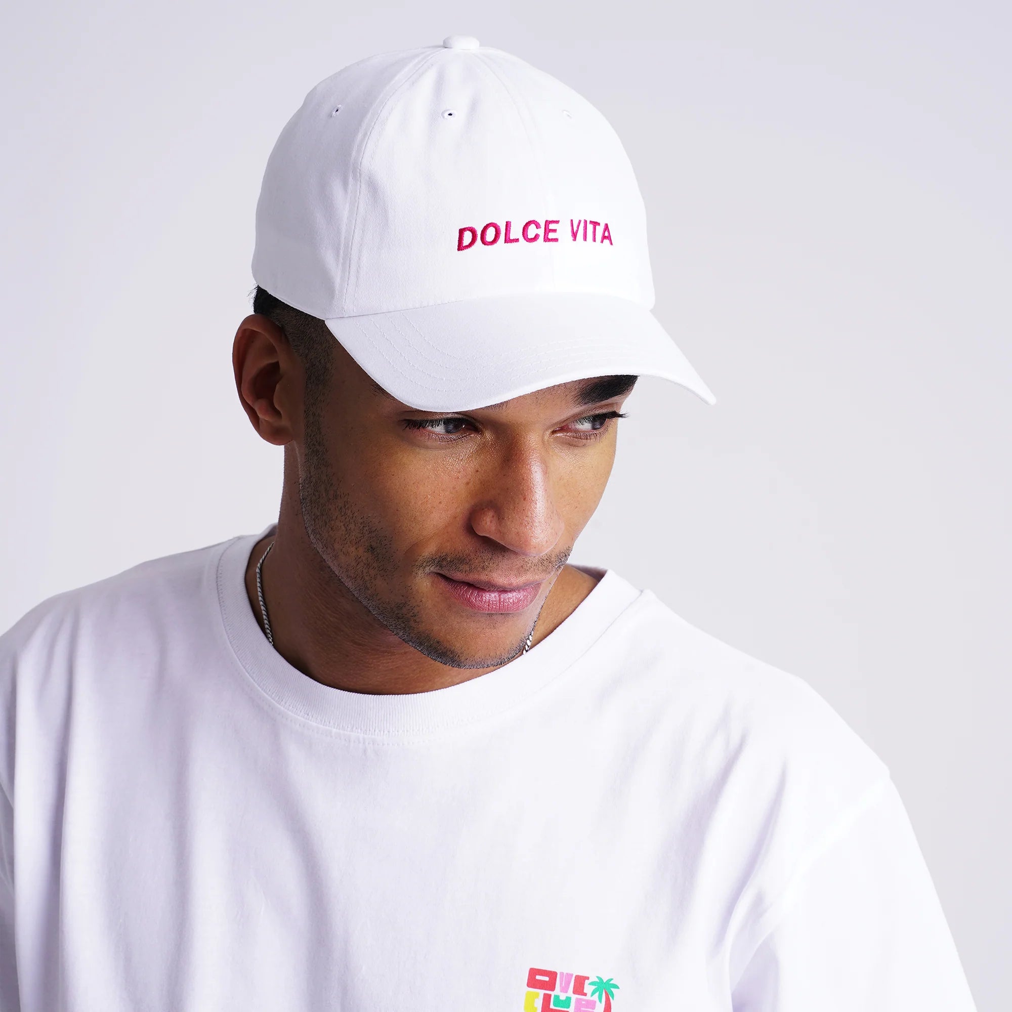 Mann trägt ein weißes Basecap mit rotem Text "DOLCE VITA" und ein weißes T-Shirt mit kleinem bunten Logo.