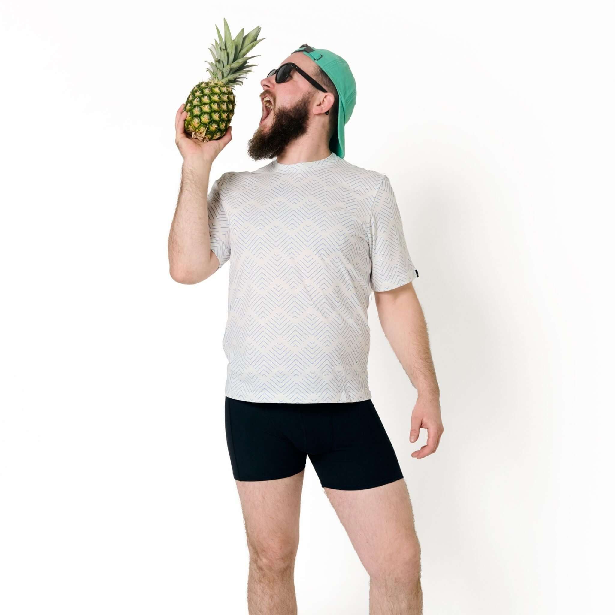Mann mit Bart und Sonnenbrille hält eine Ananas und tut so, als würde er hineinbeißen; trägt T-Shirt und Mütze rückwärts.