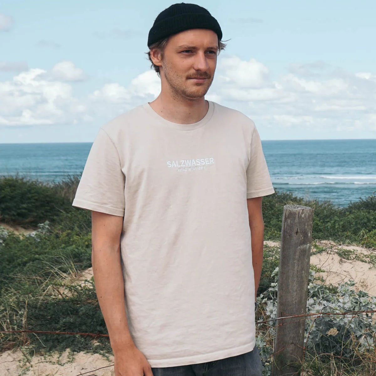 Ein Mann in einem hellbeigen T-Shirt und einer schwarzen Mütze steht im Freien in der Nähe von Sanddünen und Grünflächen, mit dem Meer und einem teilweise bewölkten Himmel im Hintergrund.