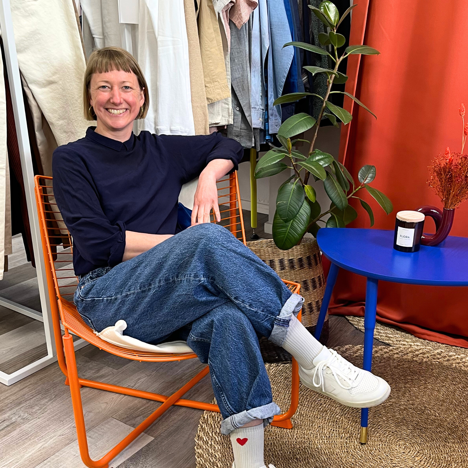 Eine lachende Person sitzt in einem Stuhl in einem Raum mit Kleidung, Pflanzen und einem kleinen Tisch.