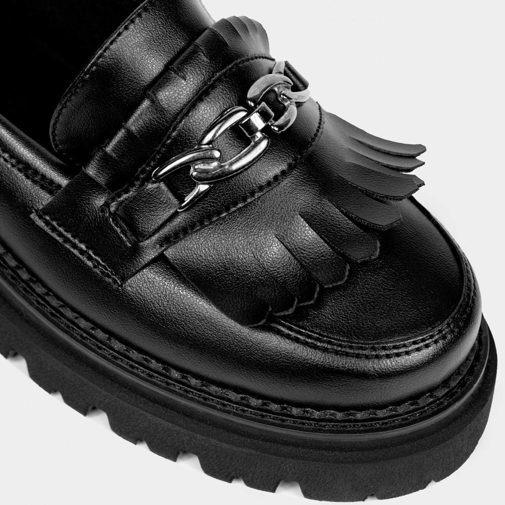 Nahaufnahme eines schwarzen Lederslippers mit dekorativen Fransen und einem silbernen Kettendetail auf dem Obermaterial, mit einer klobigen, strukturierten Sohle.