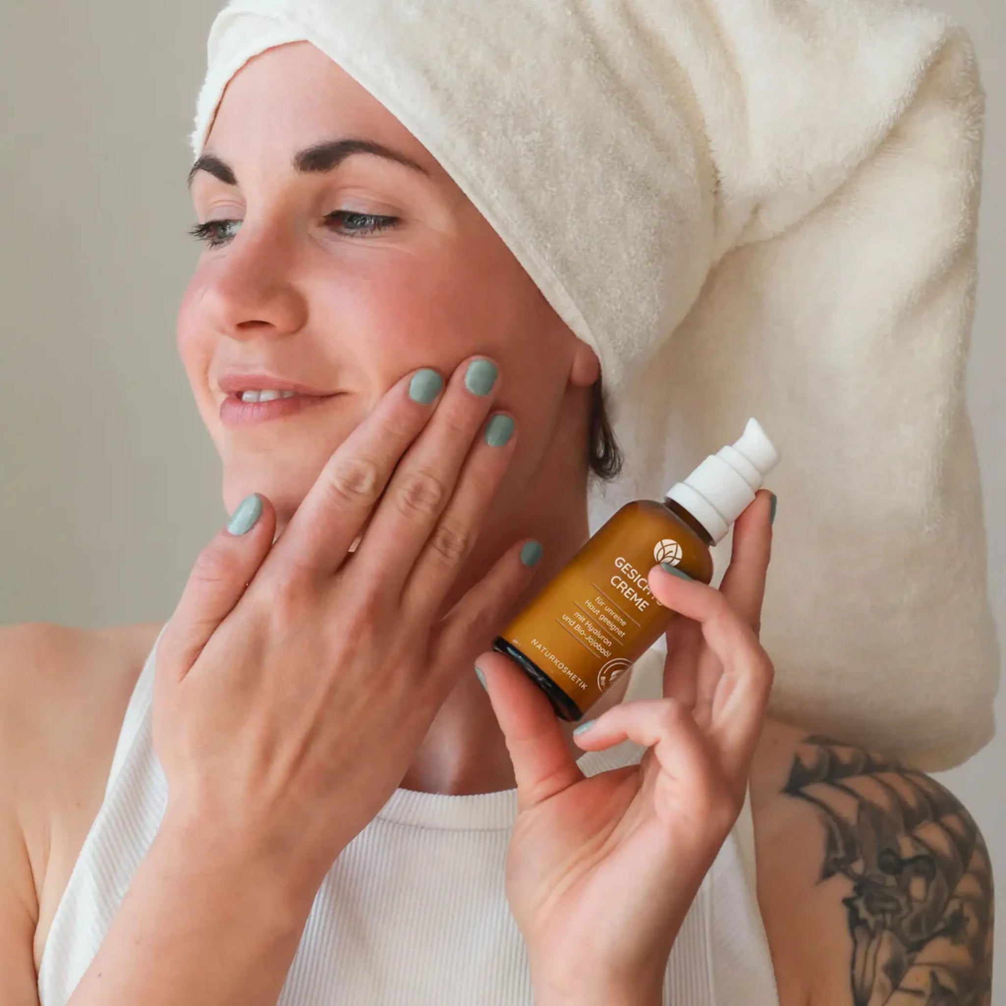 Frau mit Handtuchturban trägt Gesichtscreme auf, lächelnd hält sie eine Flasche mit der Creme in der Hand.