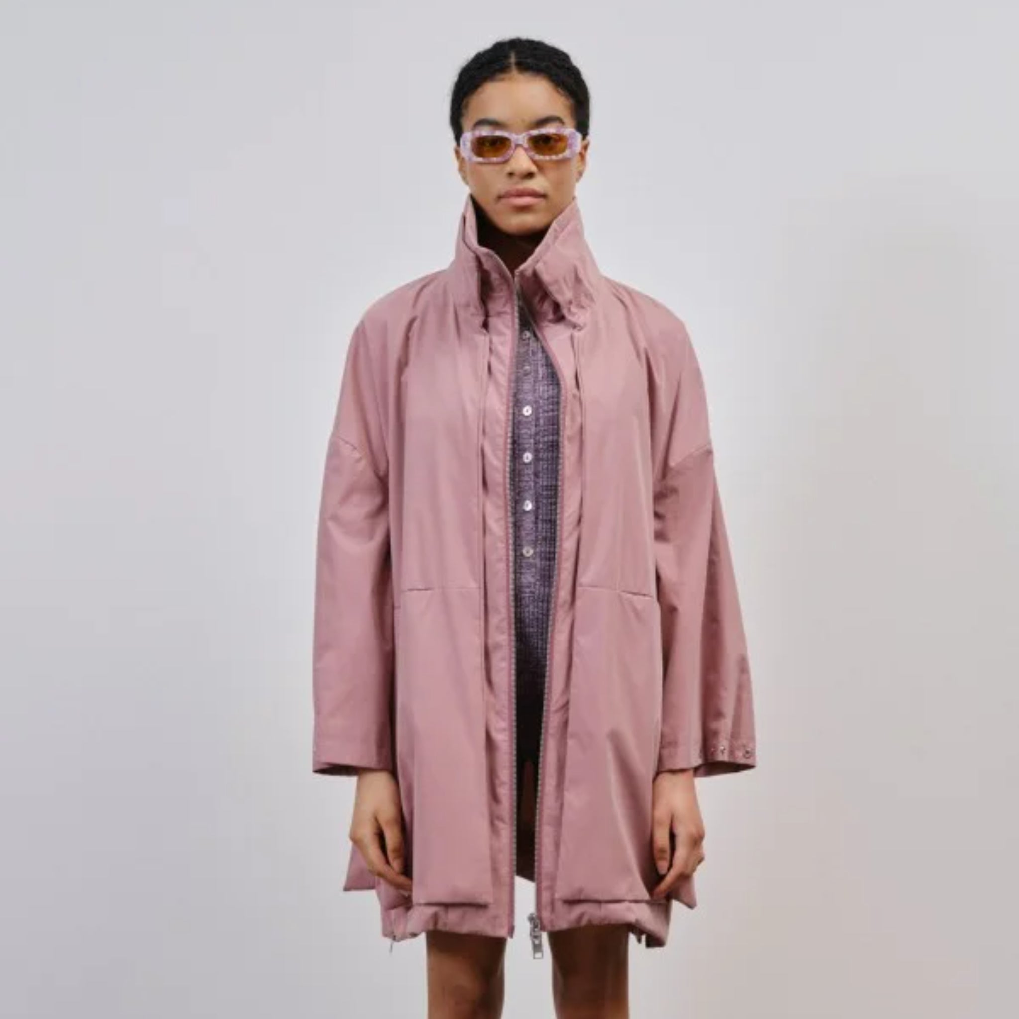 Person trägt einen rosa Mantel und eine Sonnenbrille vor einem neutralen Hintergrund.