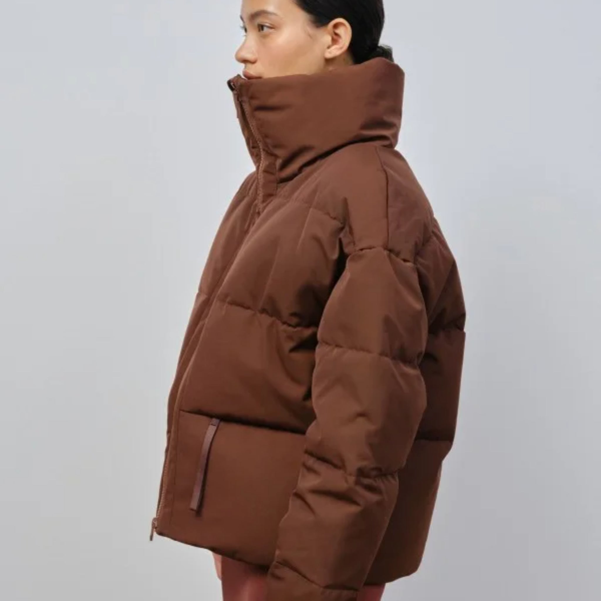 Person trägt eine braune Daunenjacke mit hohem Kragen vor neutralem Hintergrund.