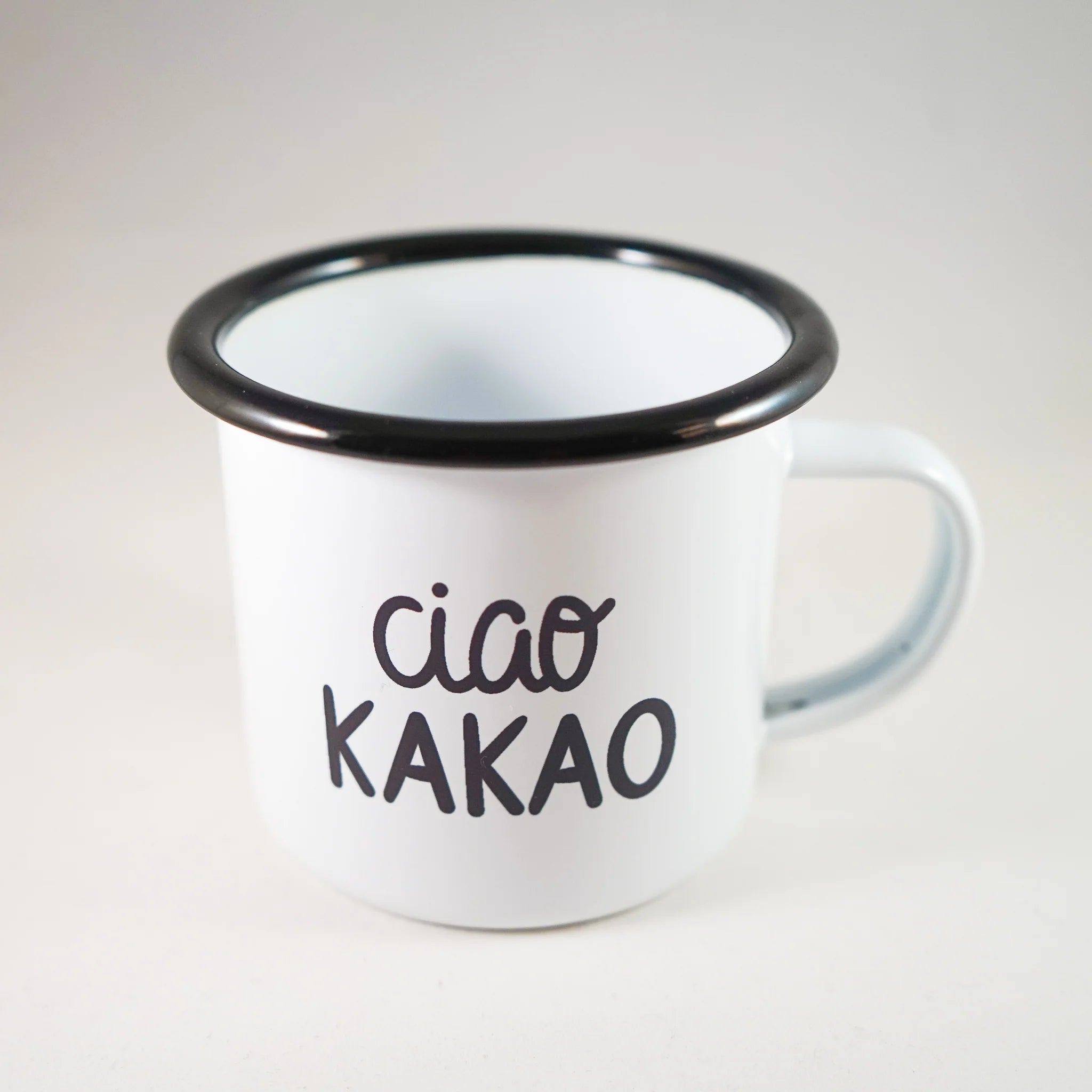Weiße Tasse mit schwarzem Rand und Aufschrift "ciao kakao" auf hellem Hintergrund.
