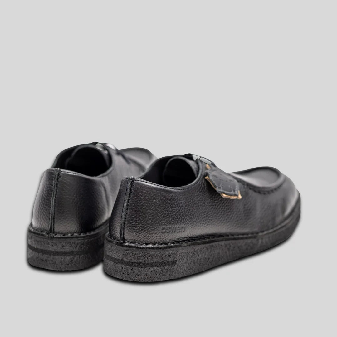 Ein Paar schwarze Lederschuhe mit Schnallenriemen, dargestellt von hinten und von der Seite, vor einem schlichten hellgrauen Hintergrund.