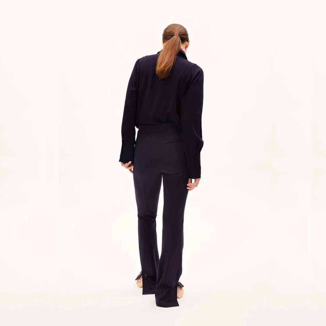 Eine Person mit zurückgebundenen langen Haaren steht mit dem Gesicht nach hinten, trägt ein schwarzes langärmeliges Hemd und eine schwarze Hose vor einem schlichten hellen Hintergrund.