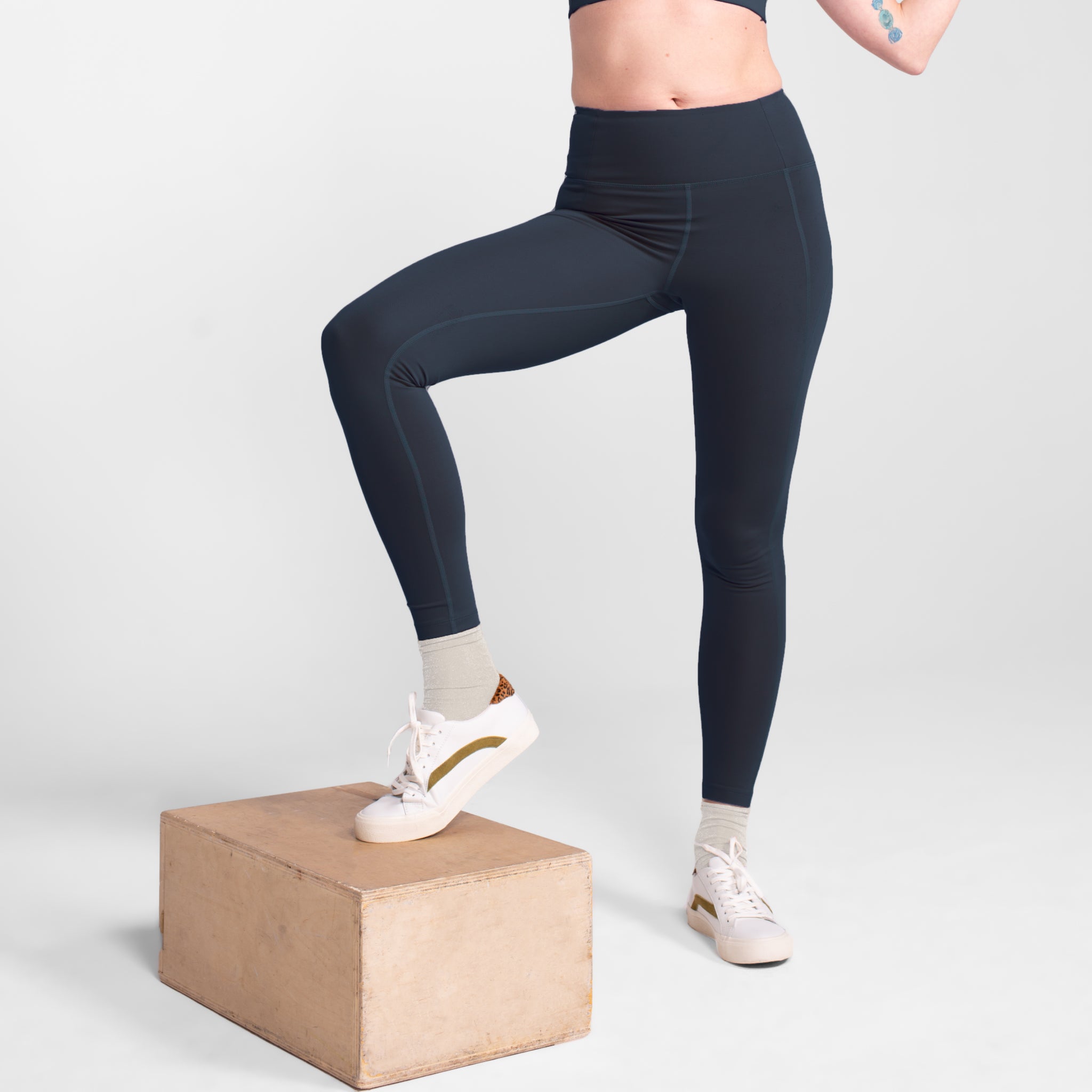 Person in schwarzen Leggings und weißen Sneakers steht mit einem Bein auf einer Box auf weißem Hintergrund.