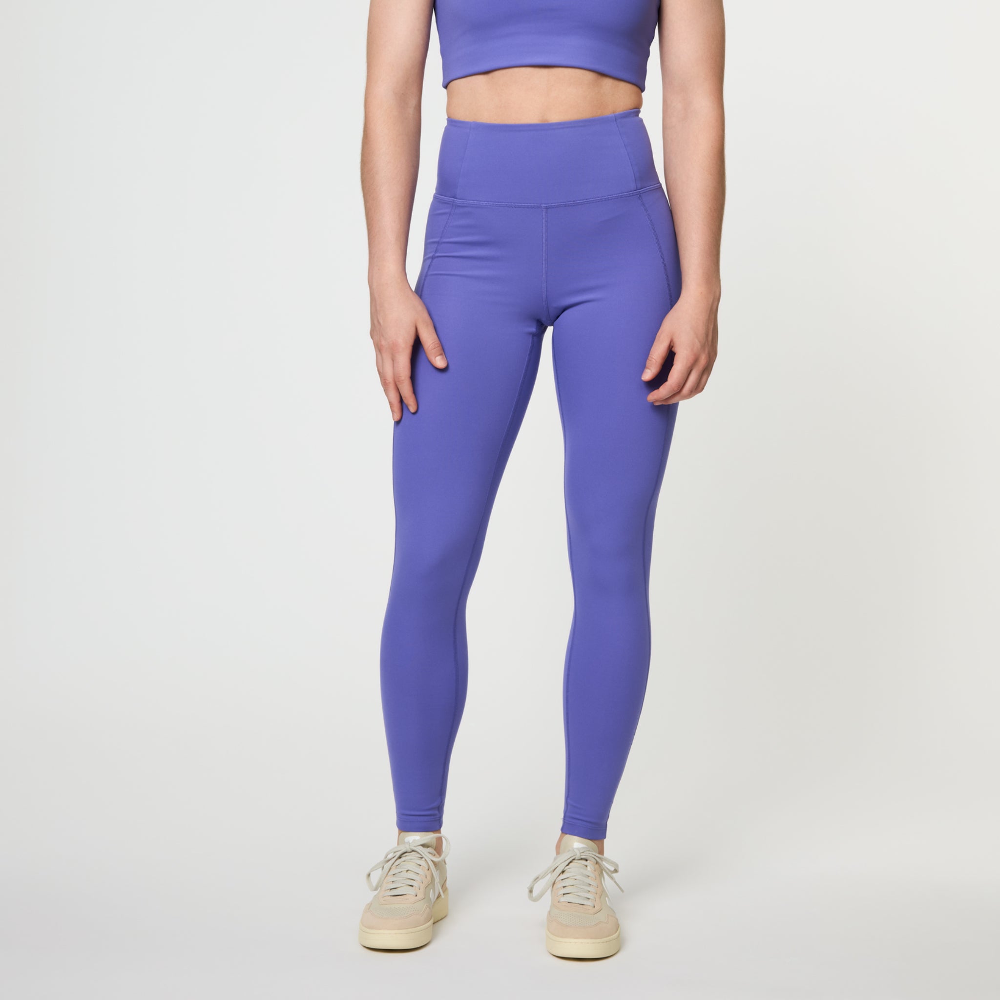 Frau trägt lila Sportleggings und ein passendes Oberteil mit hellen Sneakers vor weißem Hintergrund.