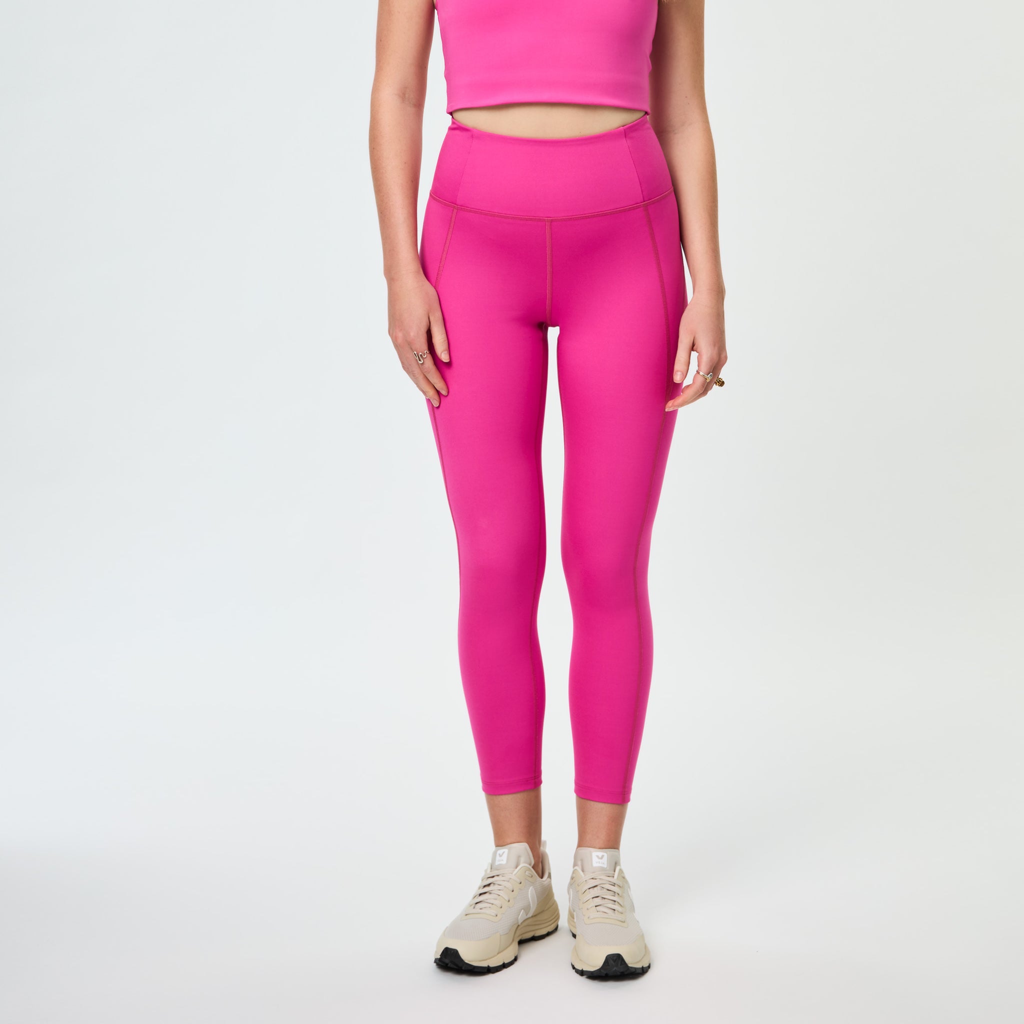 Person in pink cropped top and leggings, wearing beige sneakers, standing against a plain light background.