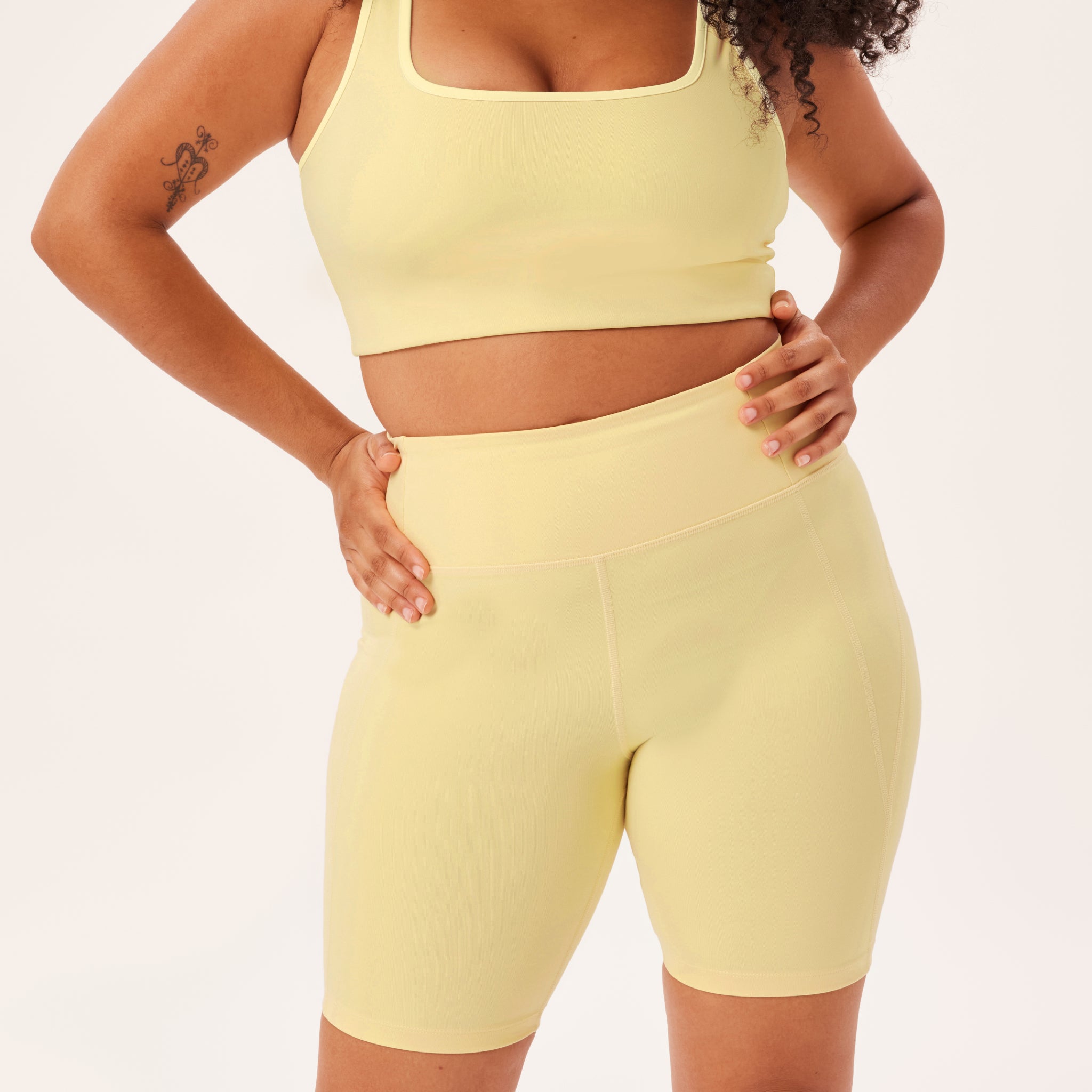 Frau trägt ein gelbes Sportoutfit mit Shorts und Top, eine Hand in die Hüfte gestützt, auf neutralem Hintergrund.