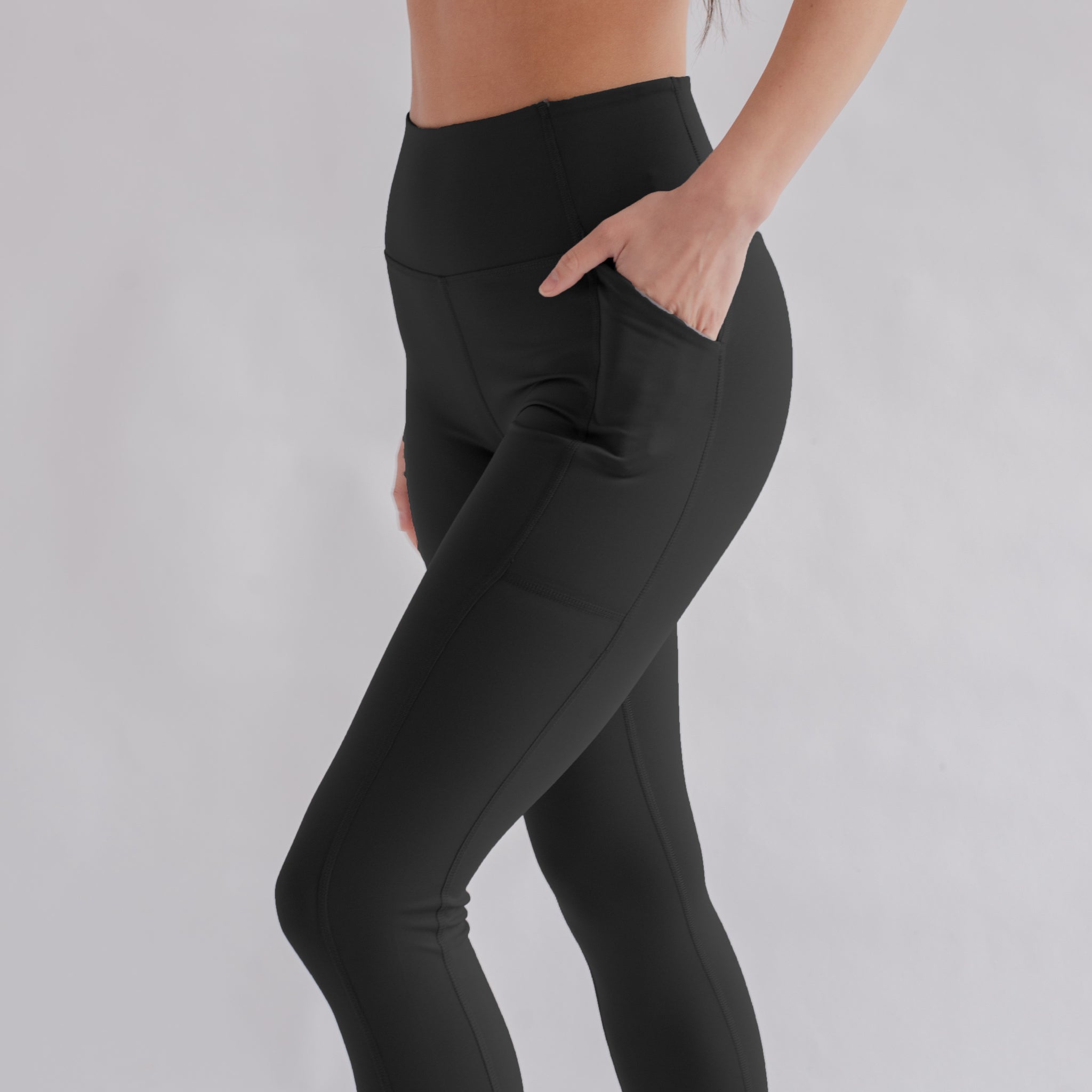 Eine Frau in schwarzer Sportleggings mit einer Hand in der Tasche, vor grauem Hintergrund.