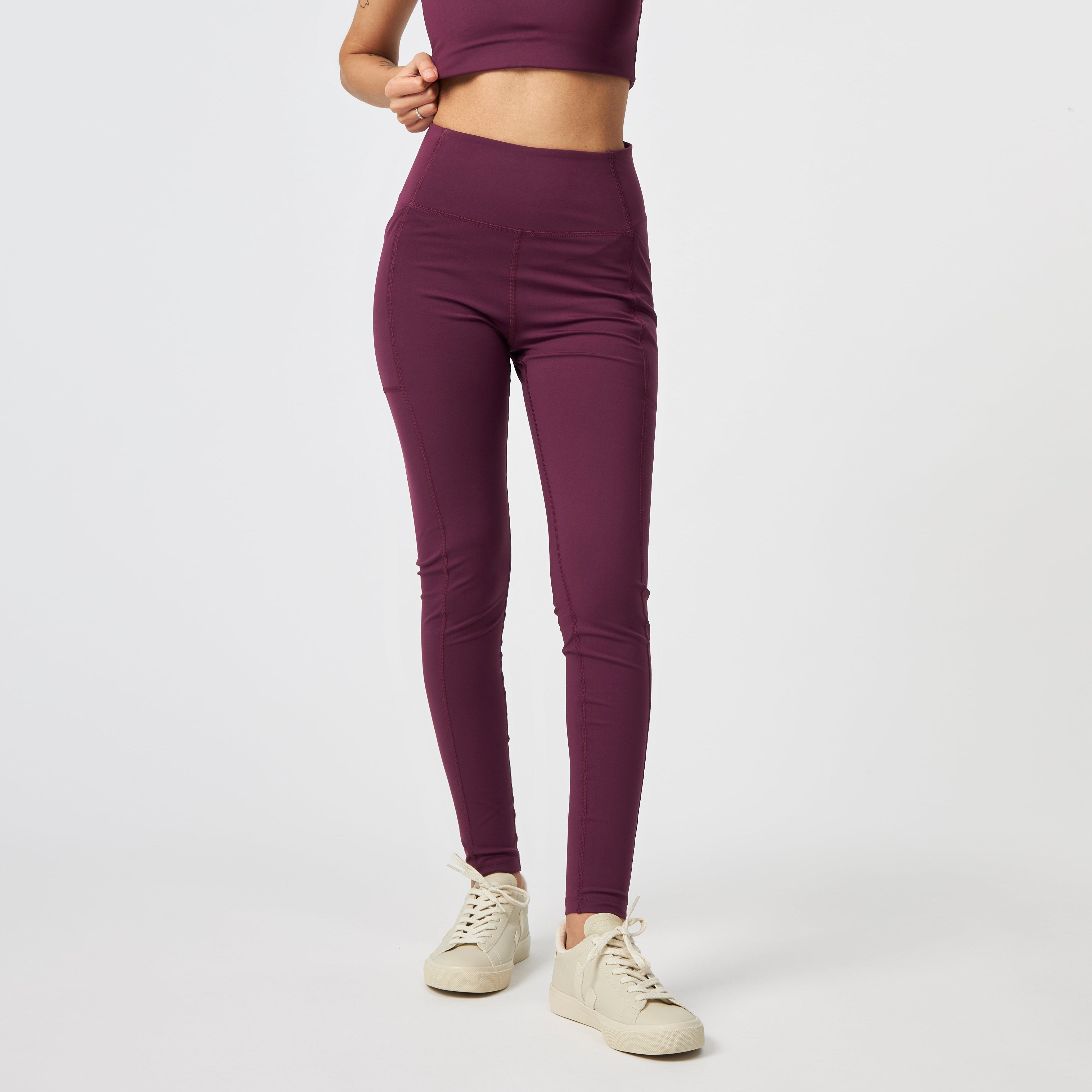 Frau in lila Leggings und Crop-Top mit beigen Sneakers posiert vor einem weißen Hintergrund.