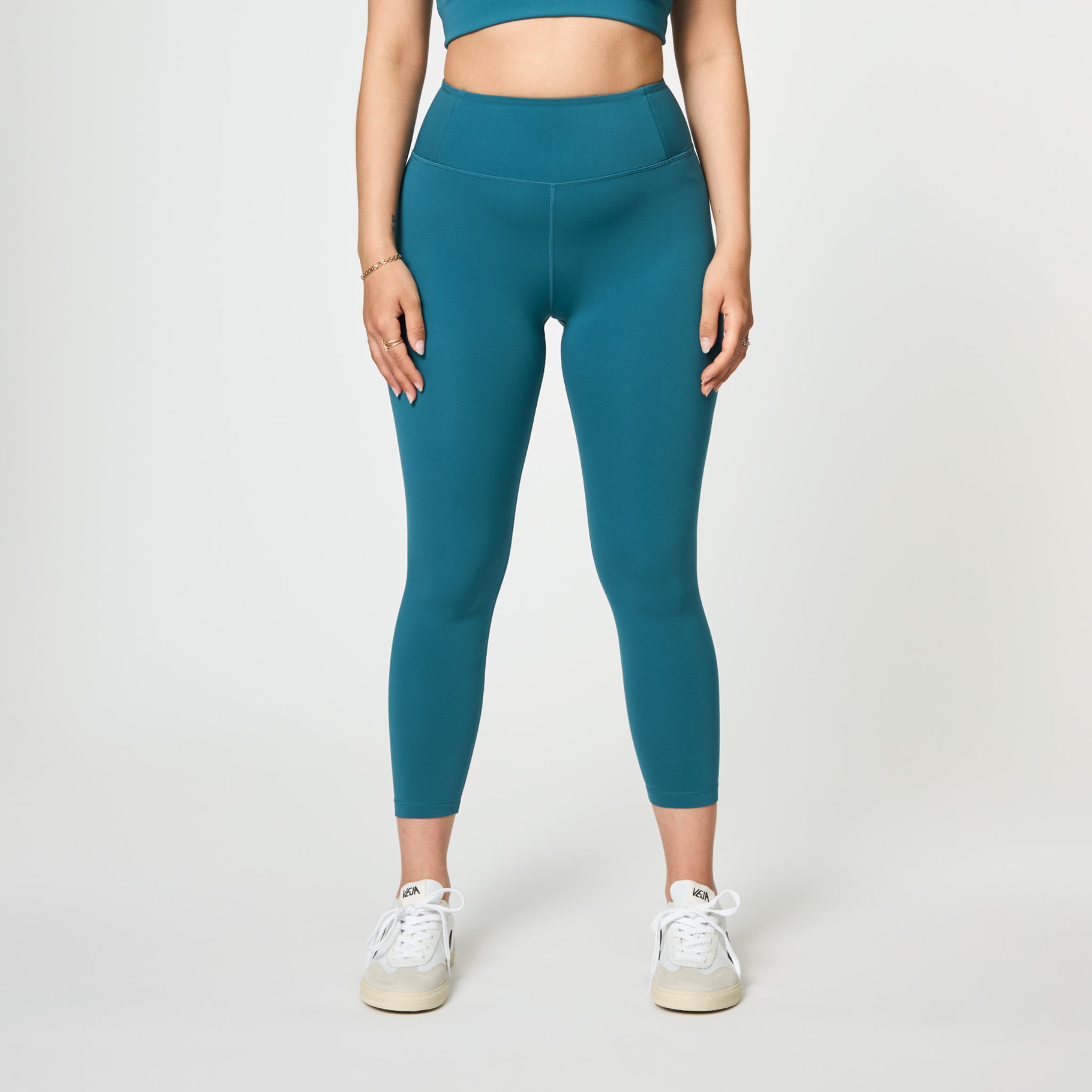 Person in teal leggings and matching crop top, wearing white sneakers, standing against a light gray background.
